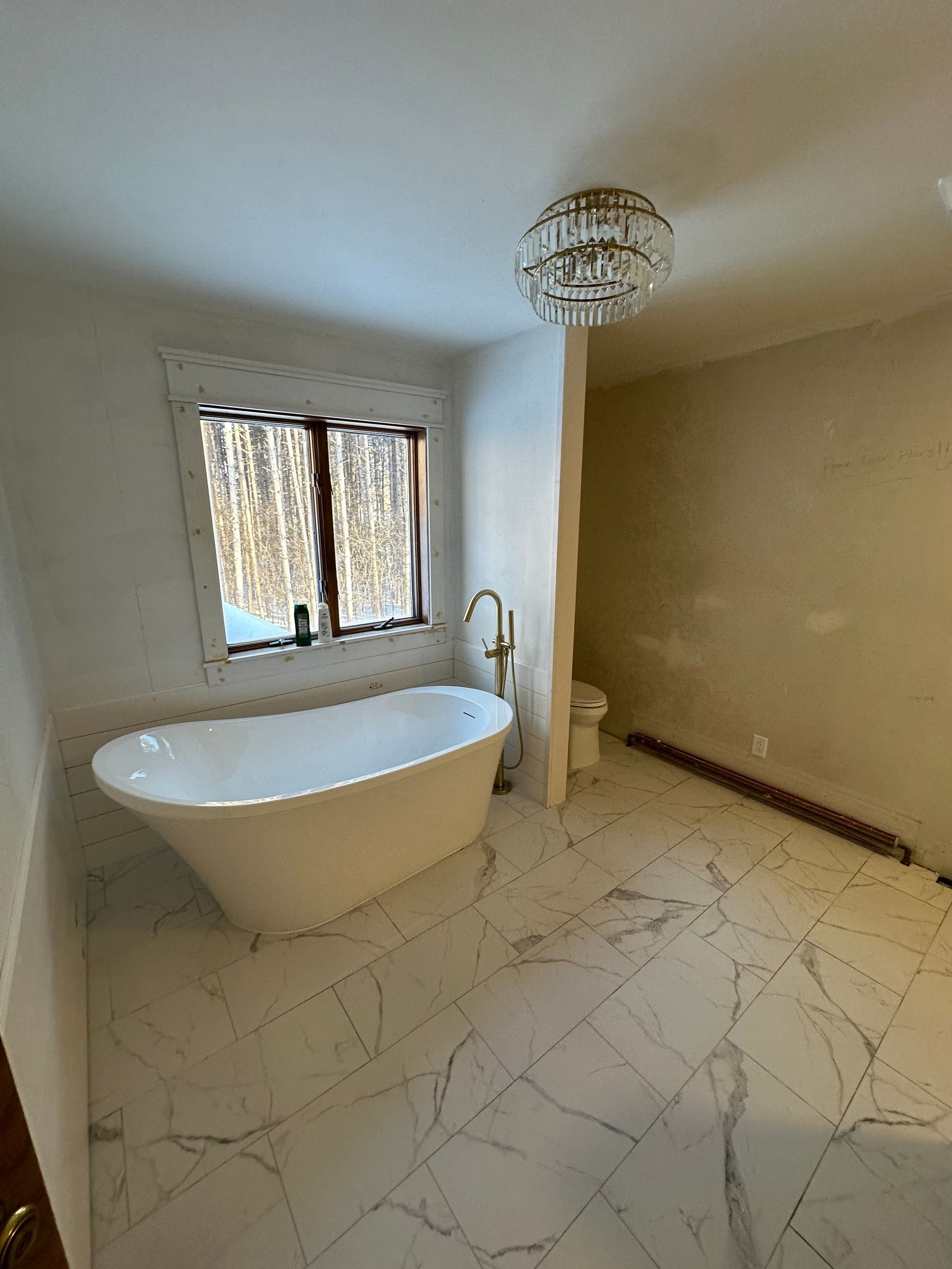 A bright, unfinished bathroom featuring a white freestanding bathtub by a window, marble-patterned tile, and a chandelier.