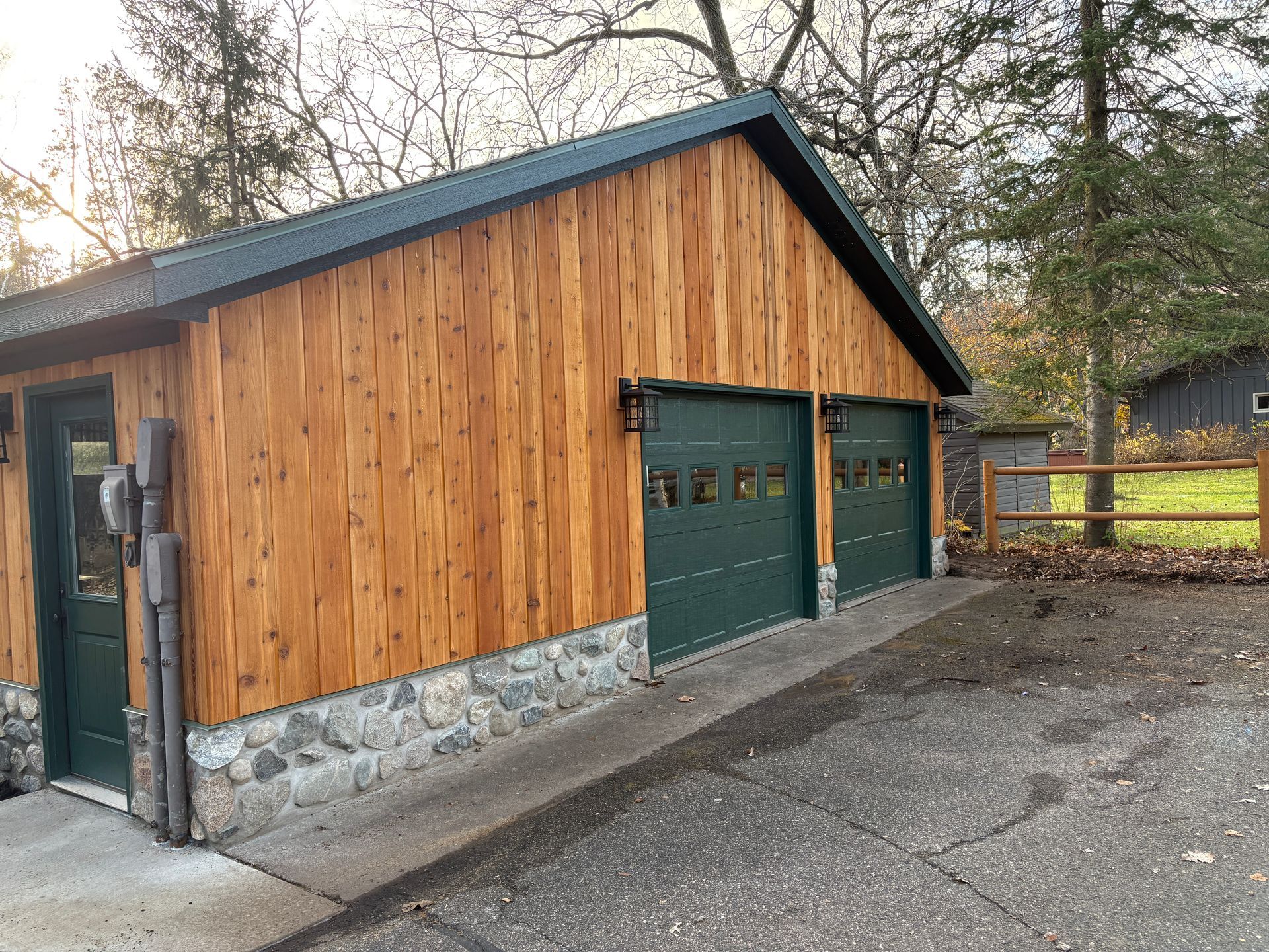 Wood-sided garage with green doors and roof. Stone base, paved driveway.