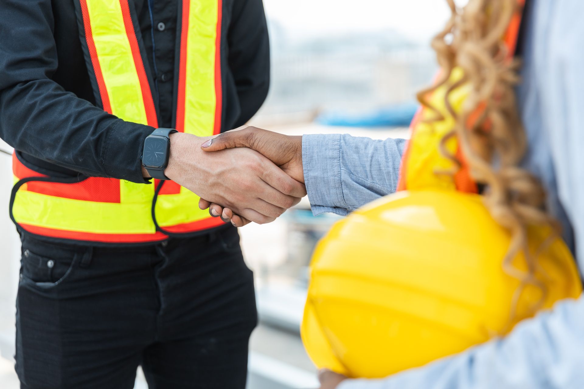 Two construction workers shaking hands, one wearing a safety vest and a hard hat is visible.