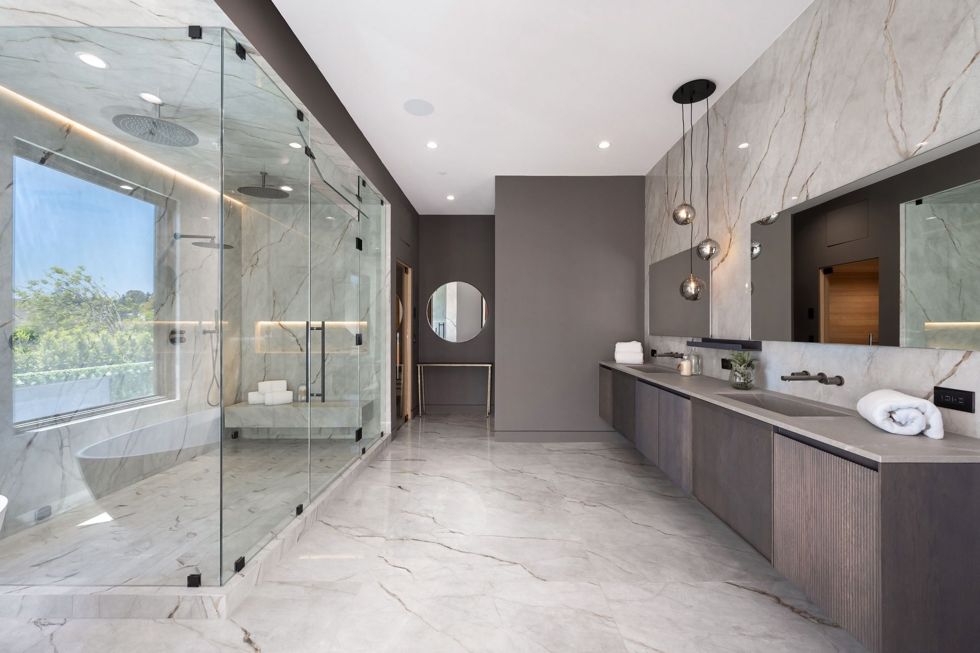 Spacious modern bathroom with glass shower, marble tile, gray cabinetry, and round mirror.