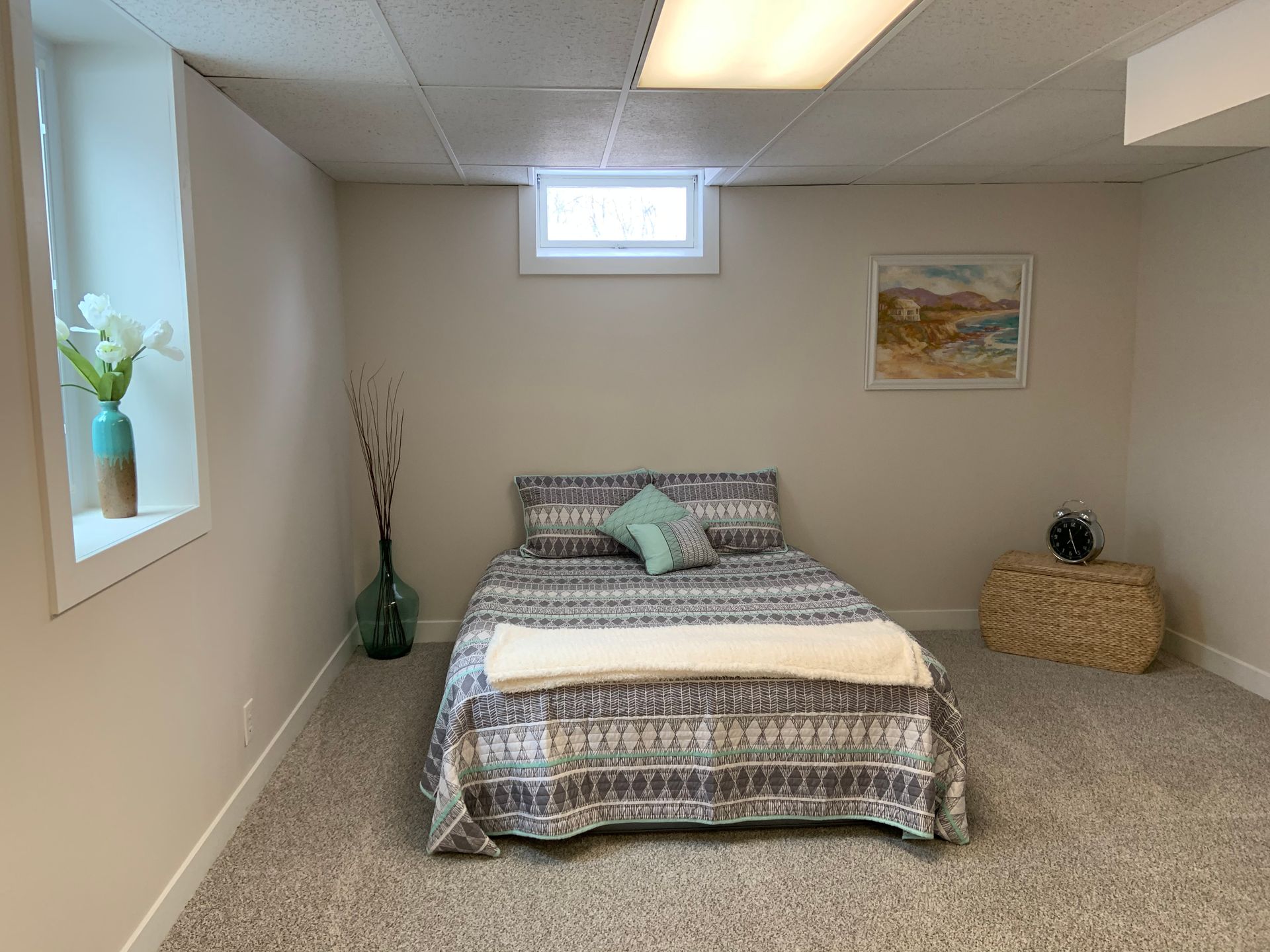 A bedroom in a basement with neutral walls, carpeted floor, a bed with a patterned comforter, and a small window.