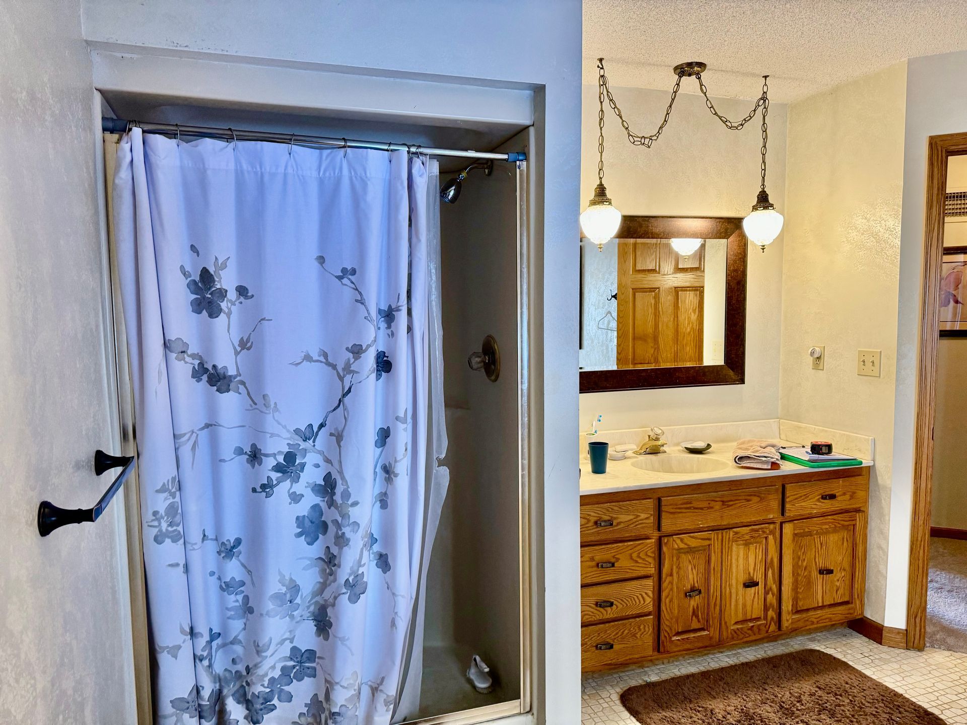 A bathroom featuring a shower with a white floral curtain beside a wooden vanity with a mirror and pendant lights.