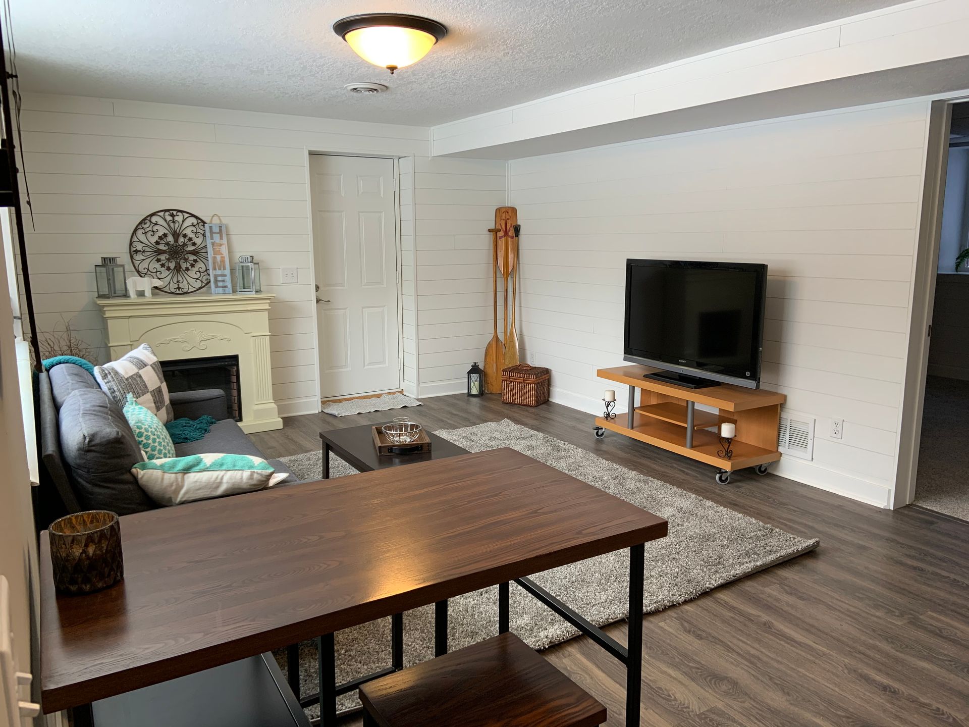A living room featuring white shiplap walls, wood-look flooring, a gray sofa, a fireplace, a TV on a stand, and a table.