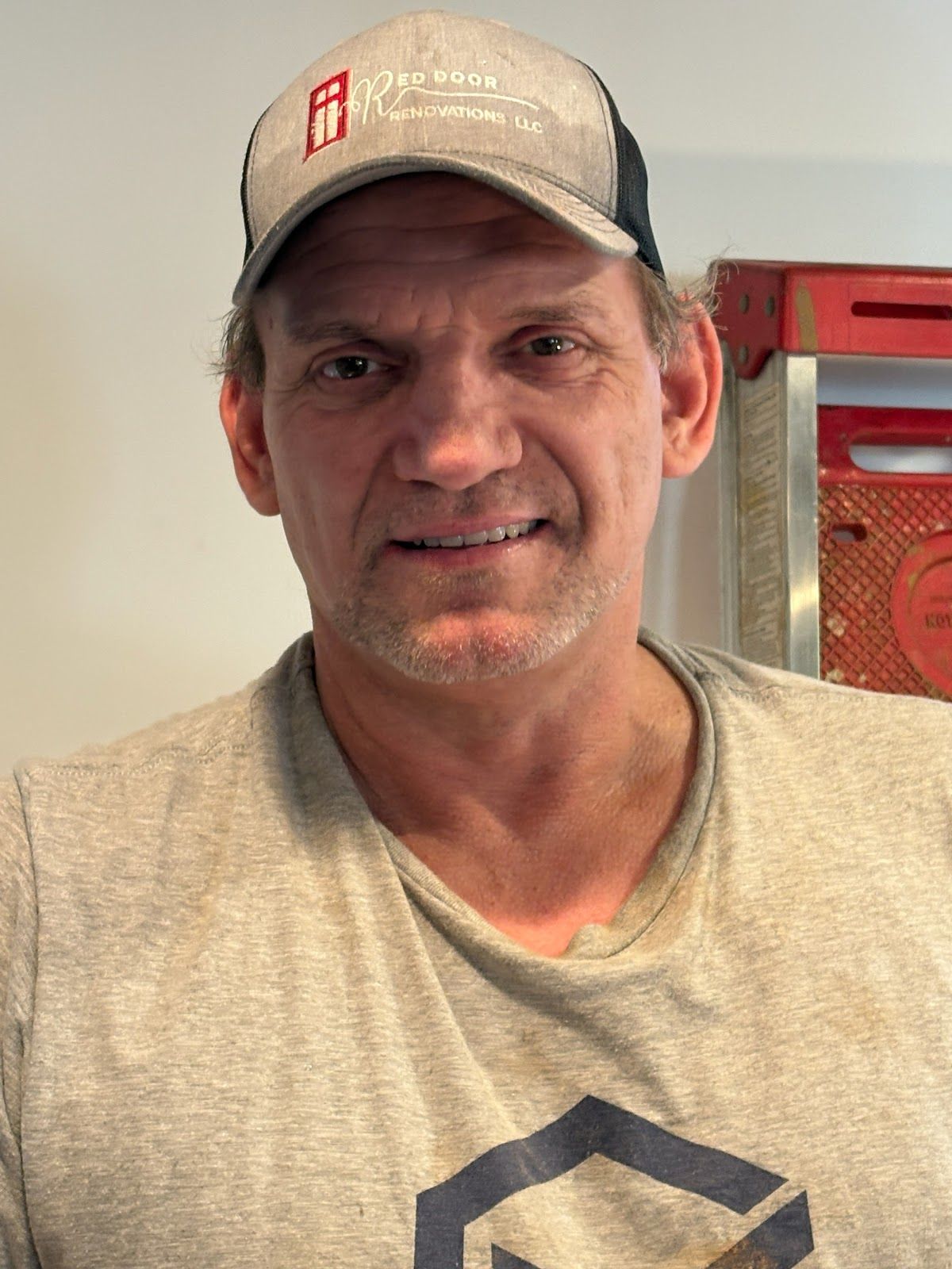 Man wearing a cap and gray t-shirt smiles at the camera, standing indoors.