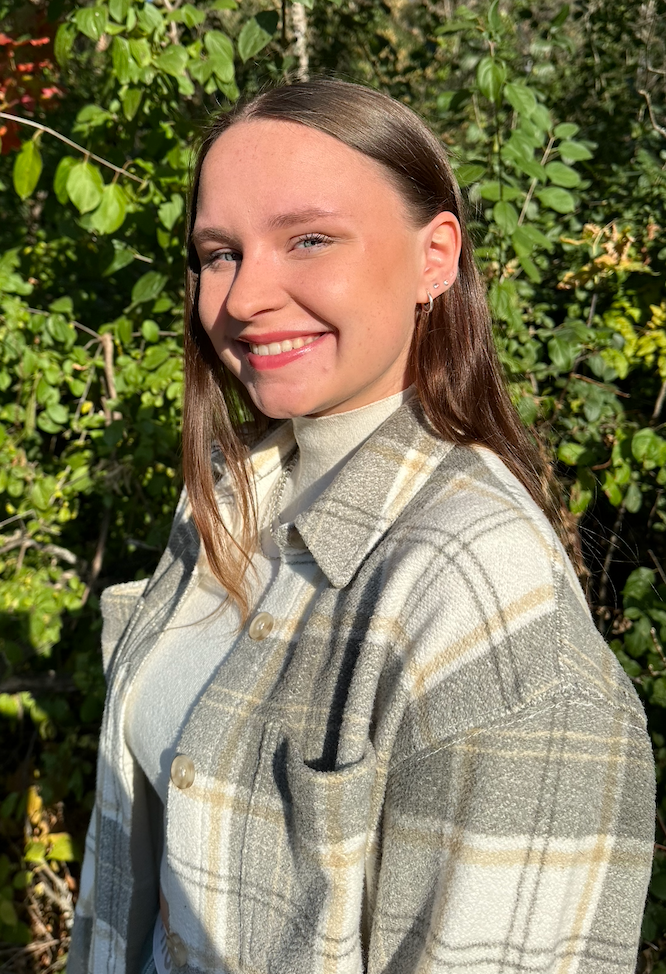 Woman smiling, wearing a white turtleneck and plaid jacket, outdoors with green foliage.