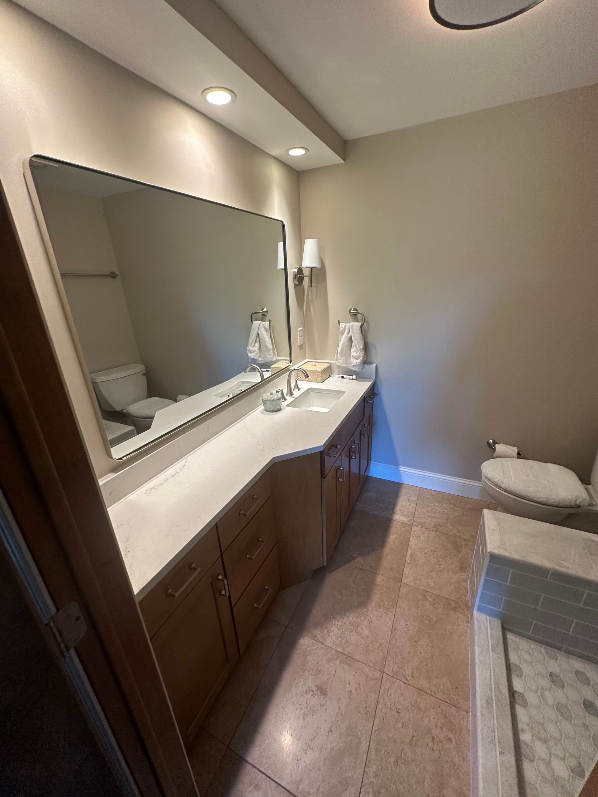 Bathroom with vanity, large mirror, and toilet. Light-colored cabinets and walls. Stone-like floor.