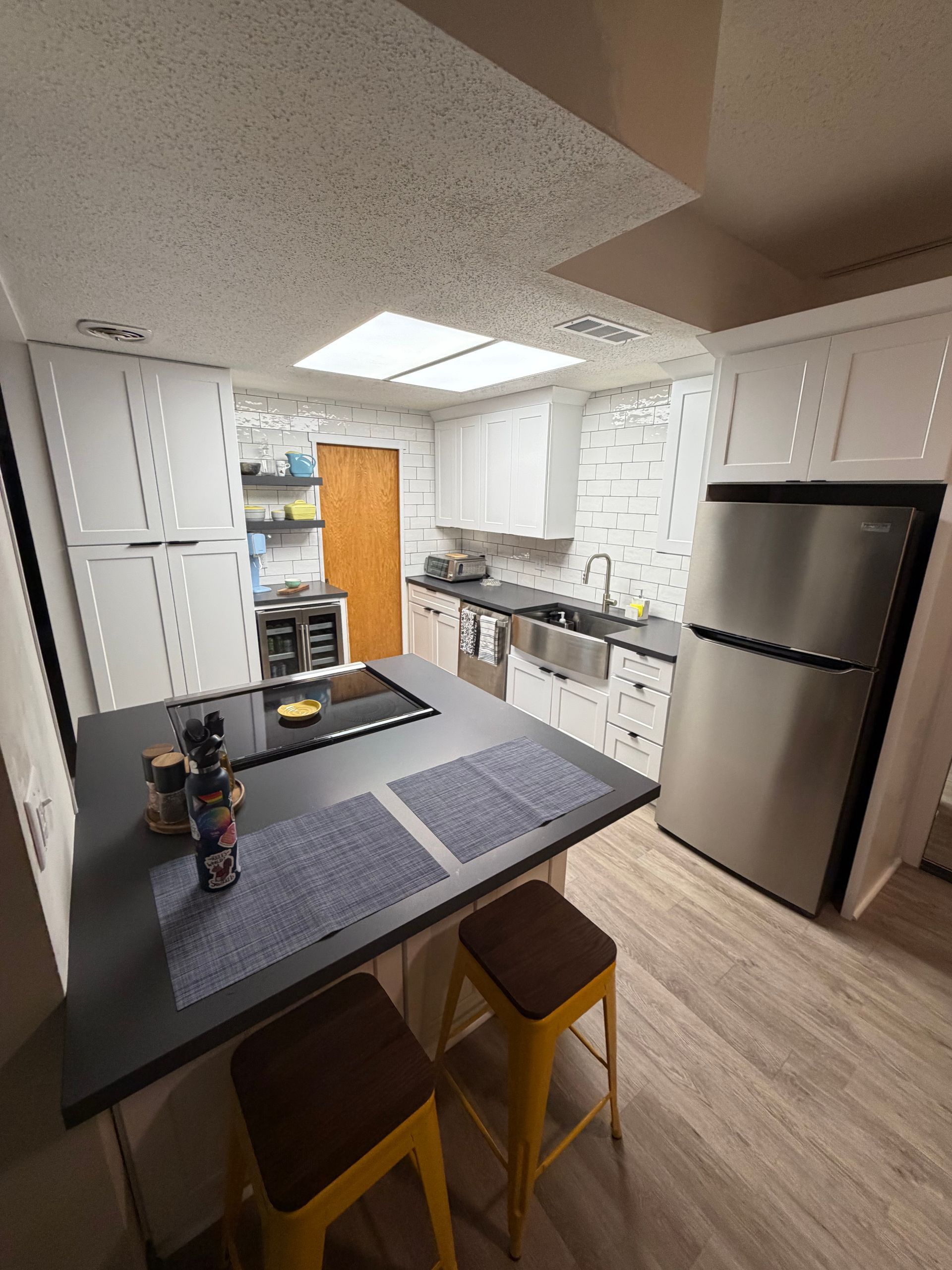 Modern kitchen with white cabinets, island with stools, stainless steel refrigerator, and skylight.