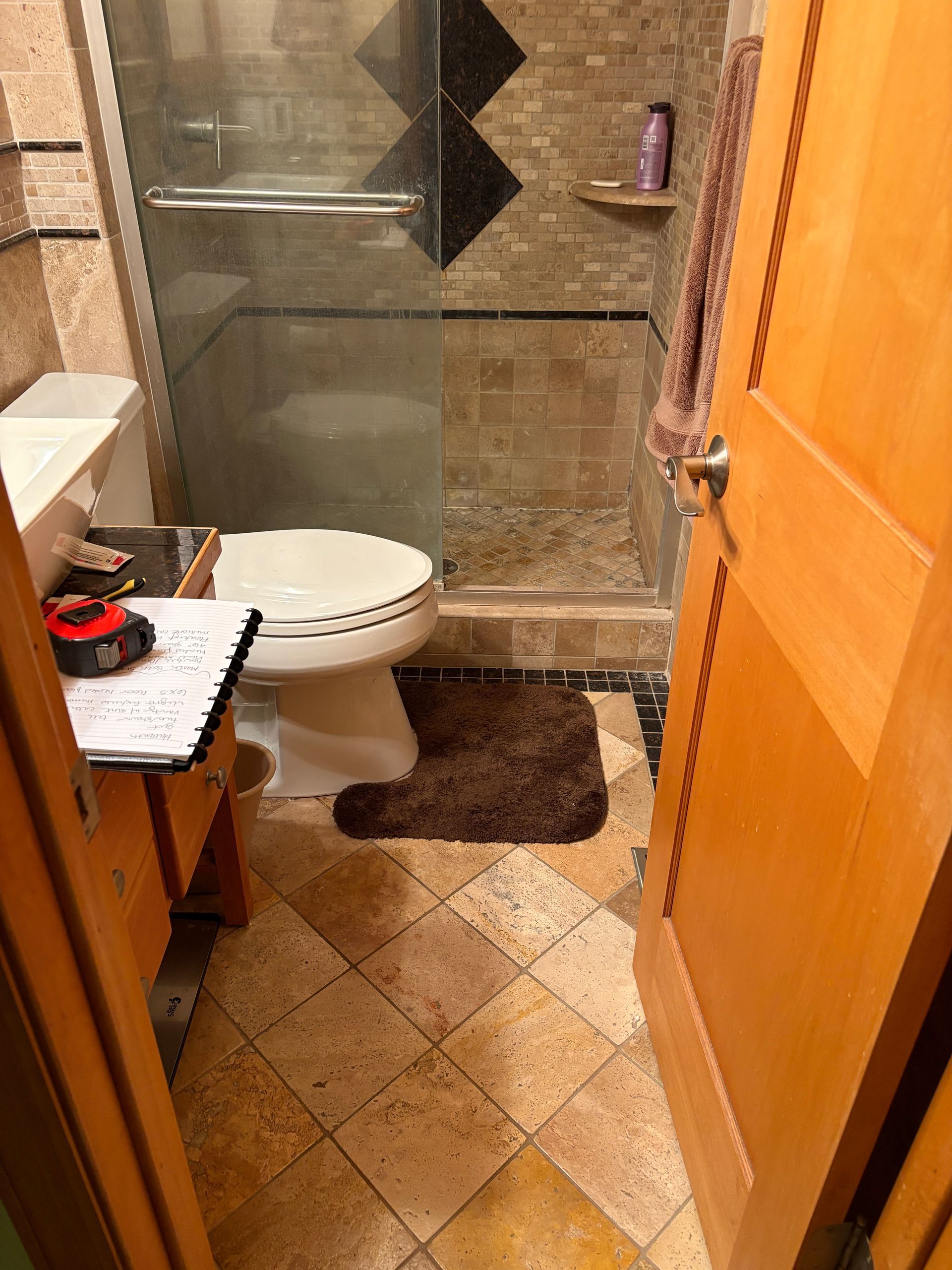 Bathroom interior: toilet, shower with glass door, brown floor rug, wood door, tiled floor.