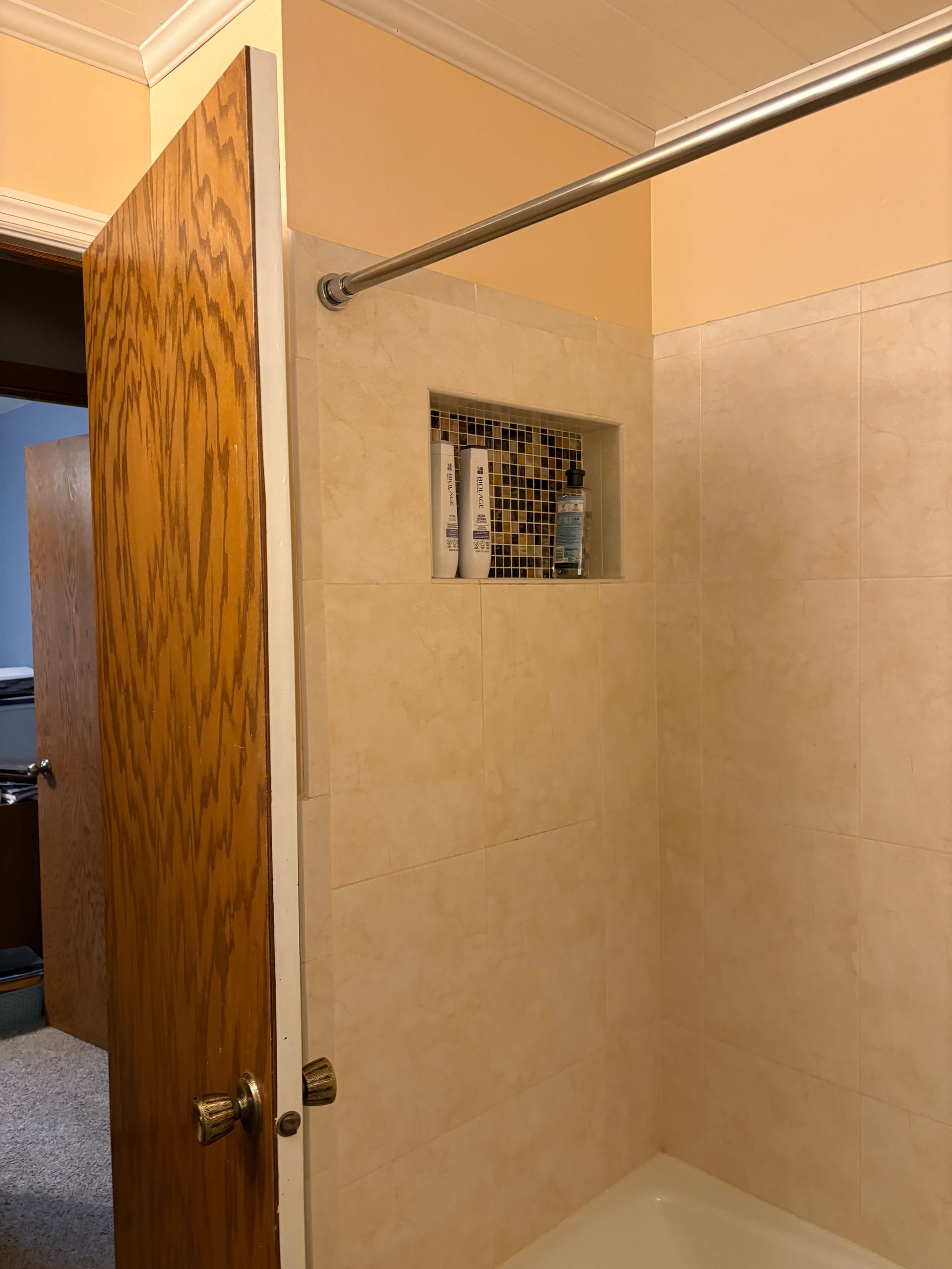 Shower with tile walls, built-in shelf, and a shower rod. A door is visible to the left.