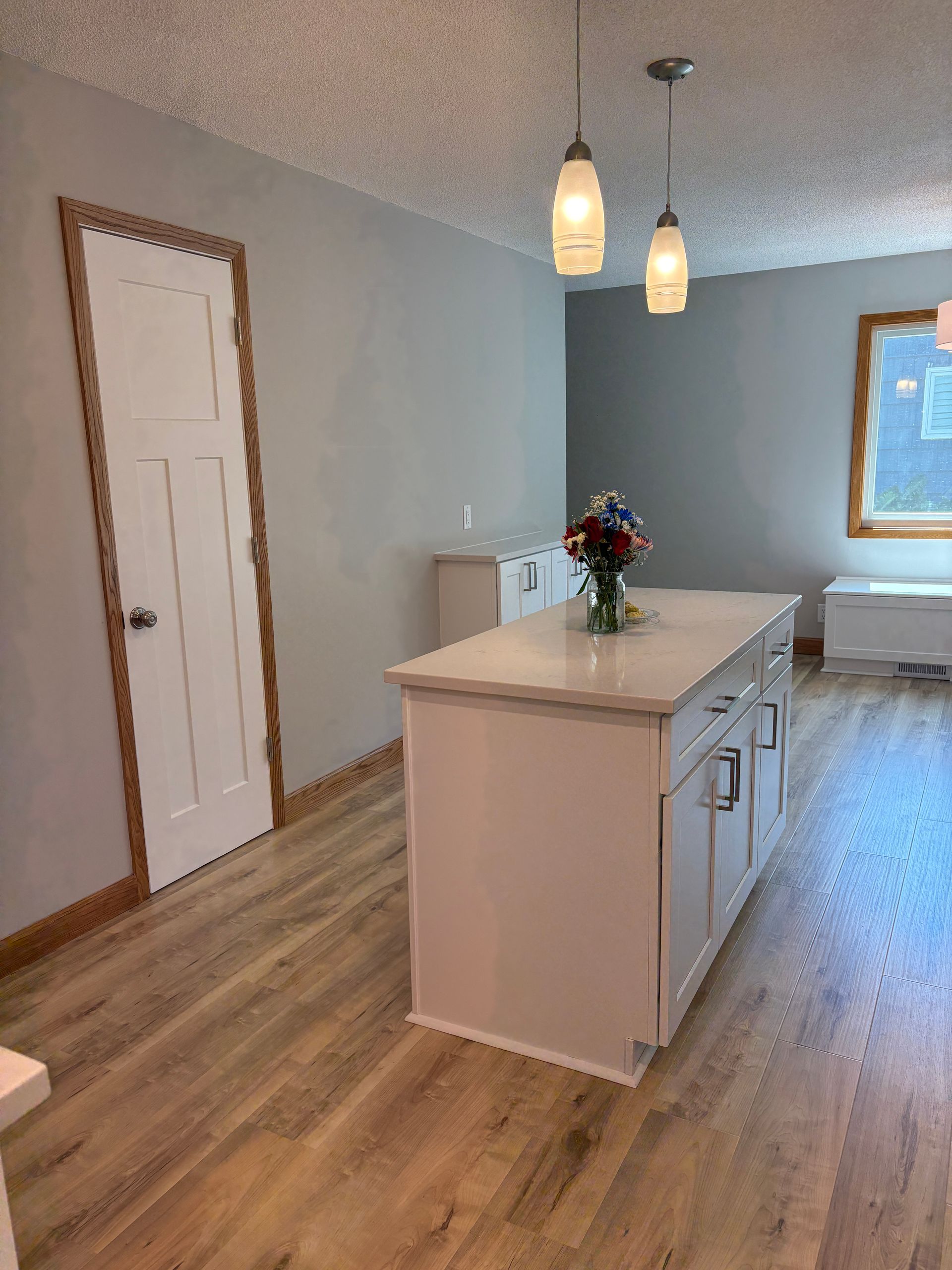 White kitchen island with flowers, pendant lights, and wooden floors. Light blue walls and white cabinets.