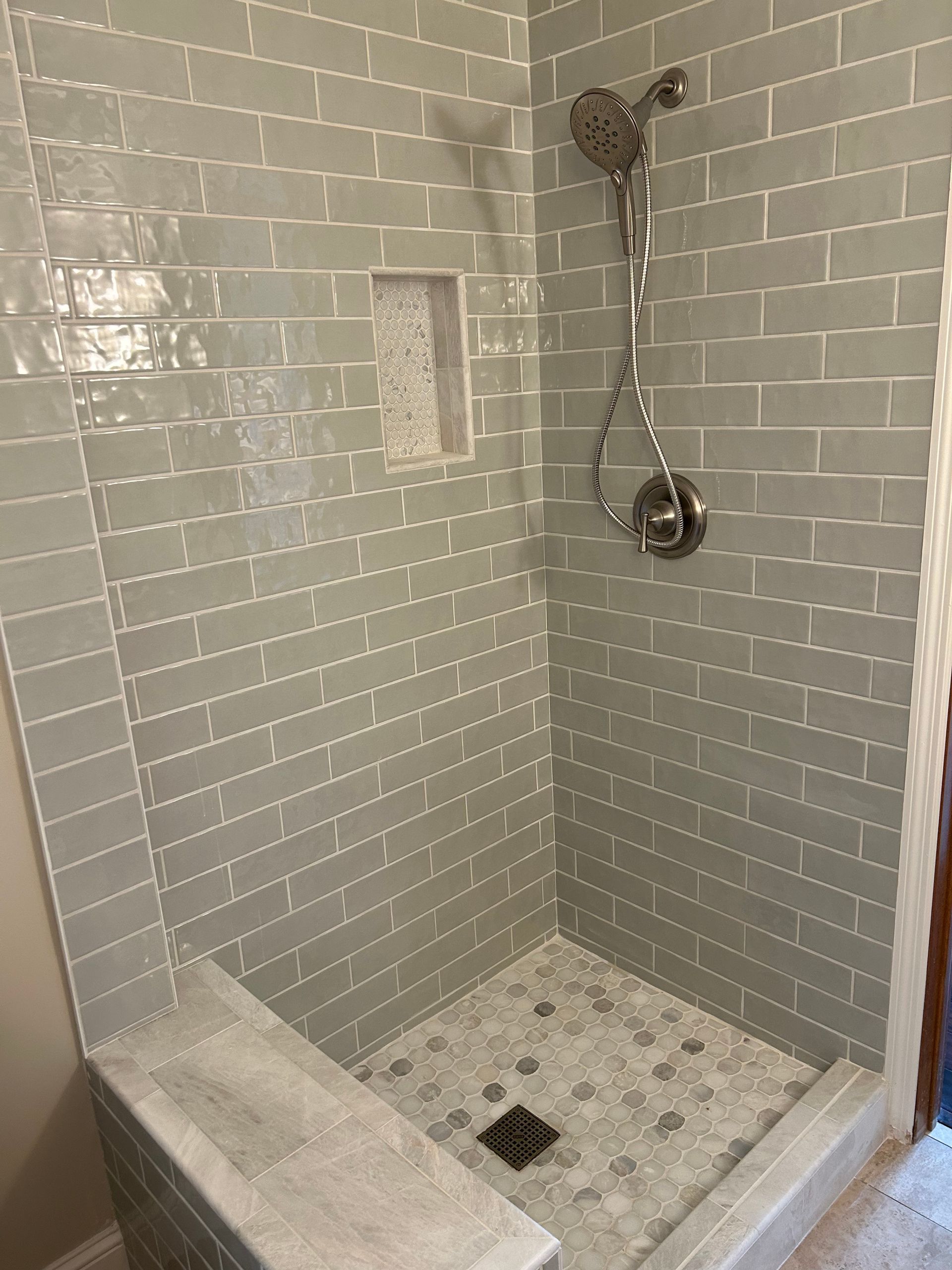 Shower with light gray subway tile walls and mosaic floor, built-in bench, and silver shower head.