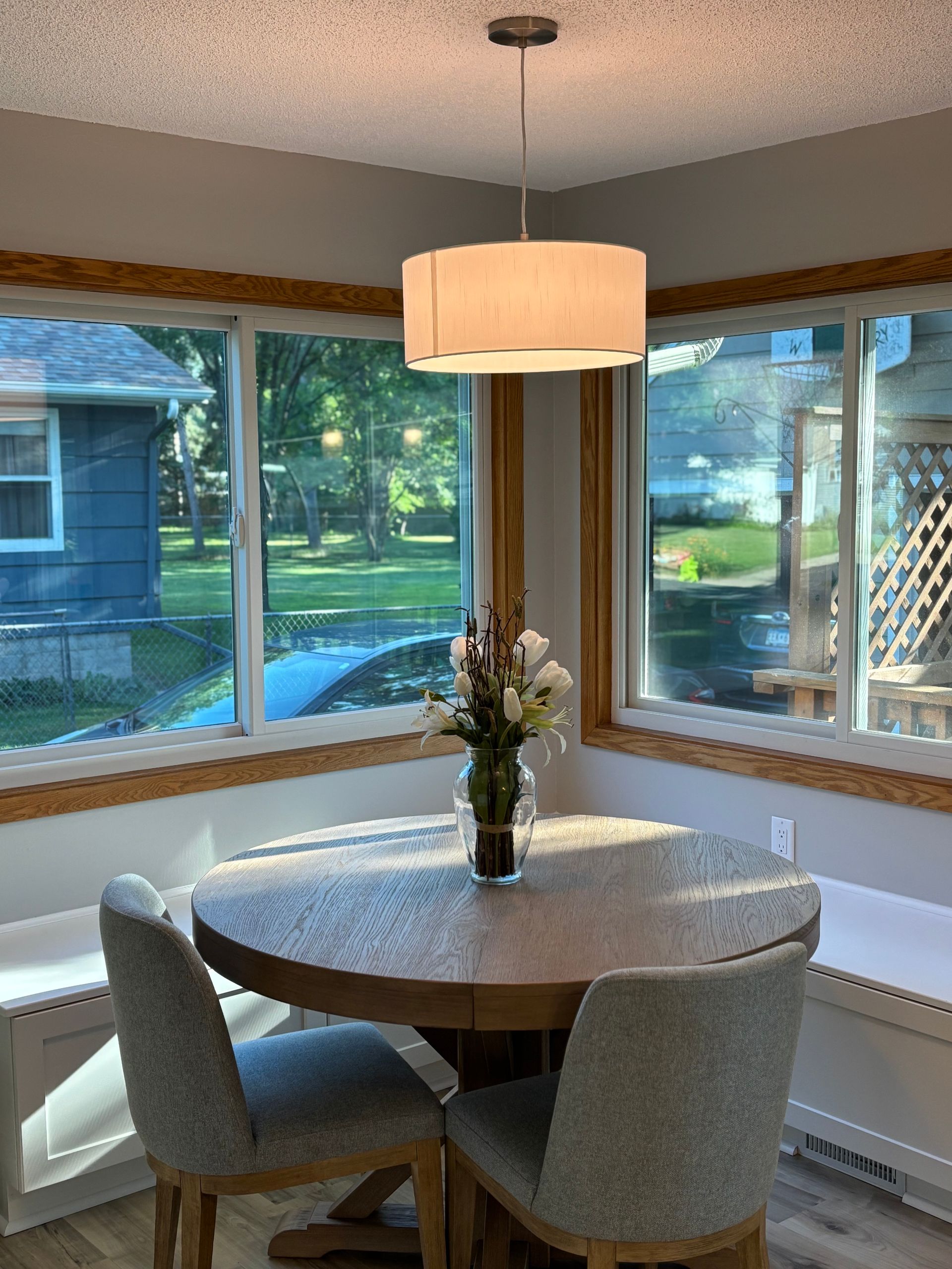 Dining area with round table, two chairs, and a hanging light. Large windows offer a view of a backyard.