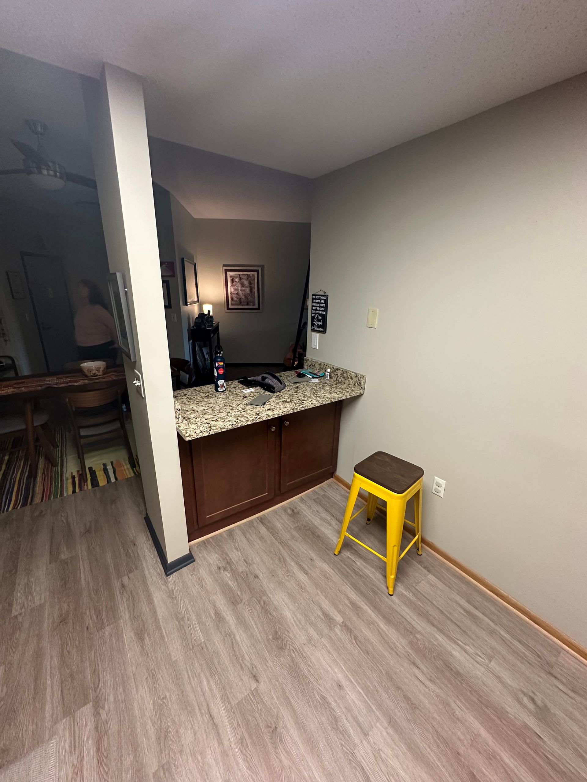 A small kitchen counter with granite top and brown cabinets, a yellow stool, and a door to the left.