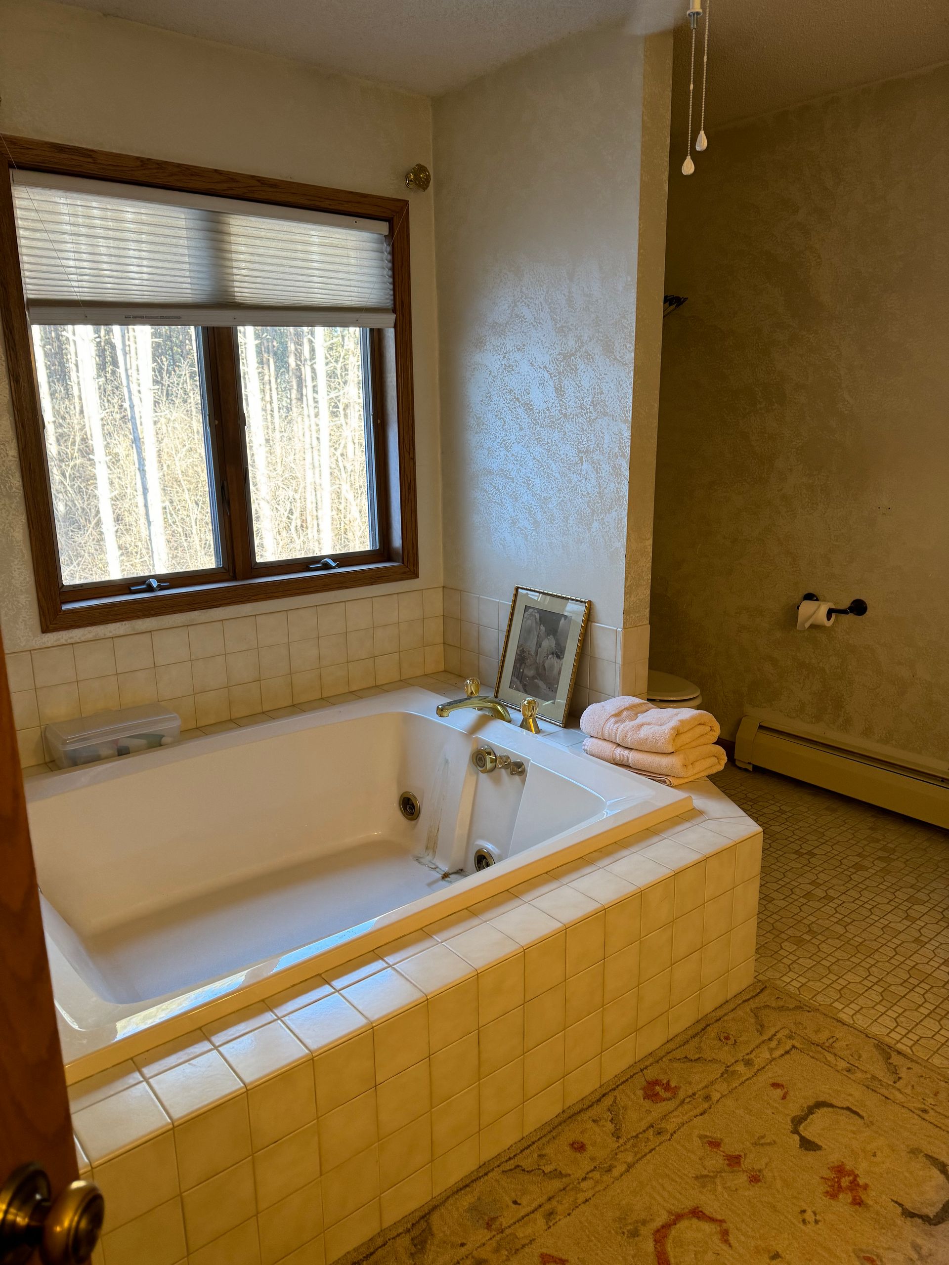 Bathroom with a white built-in tub, window, and beige tiled surround. Soft towels sit next to the tub.