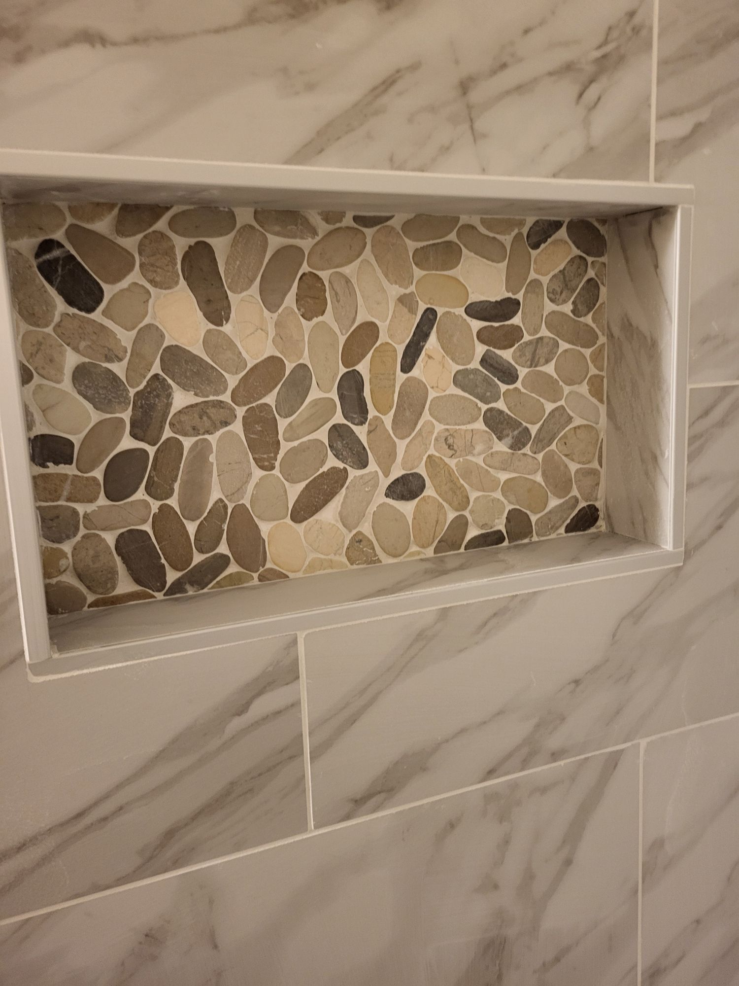 Built-in shower niche with pebble tile backing, framed by white tile and marble-patterned wall tiles.