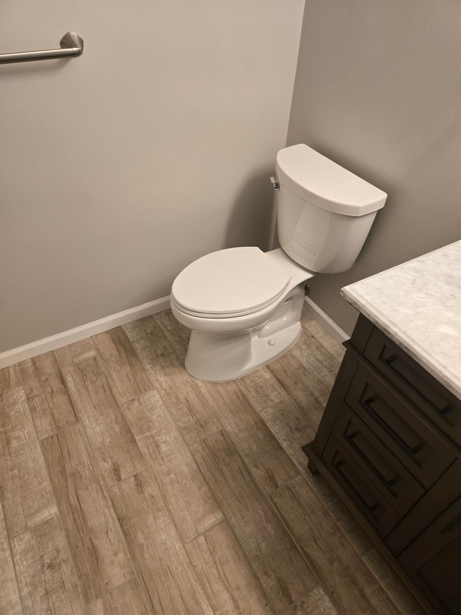 A white toilet in a bathroom with wood-look flooring and a gray wall. A dark brown vanity is on the right.