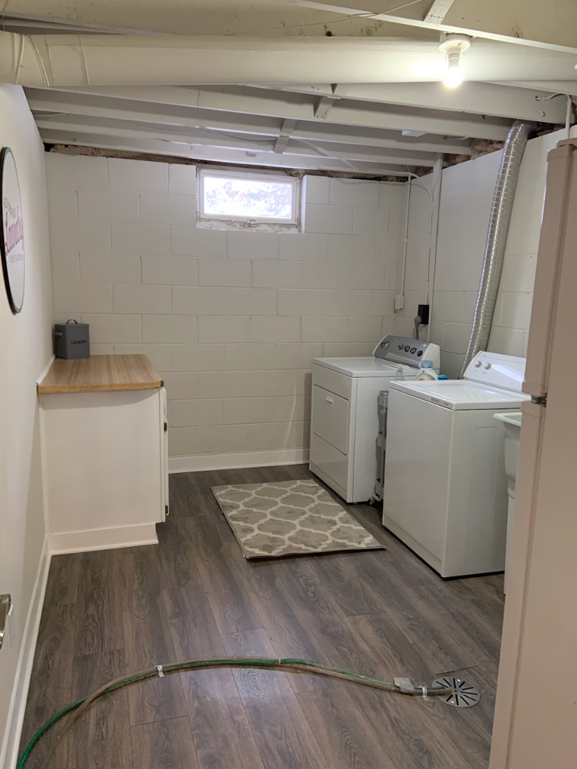 Basement laundry room with white appliances, gray rug, and wood-look flooring. A cabinet and hose are visible.
