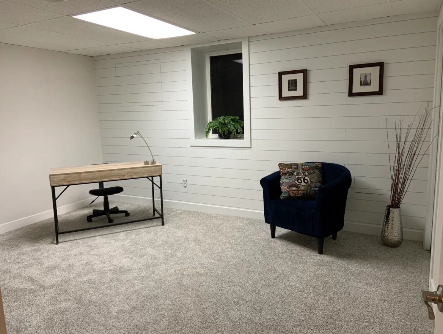Office space with a desk, chair, blue armchair, window, and white shiplap walls. Gray carpet.