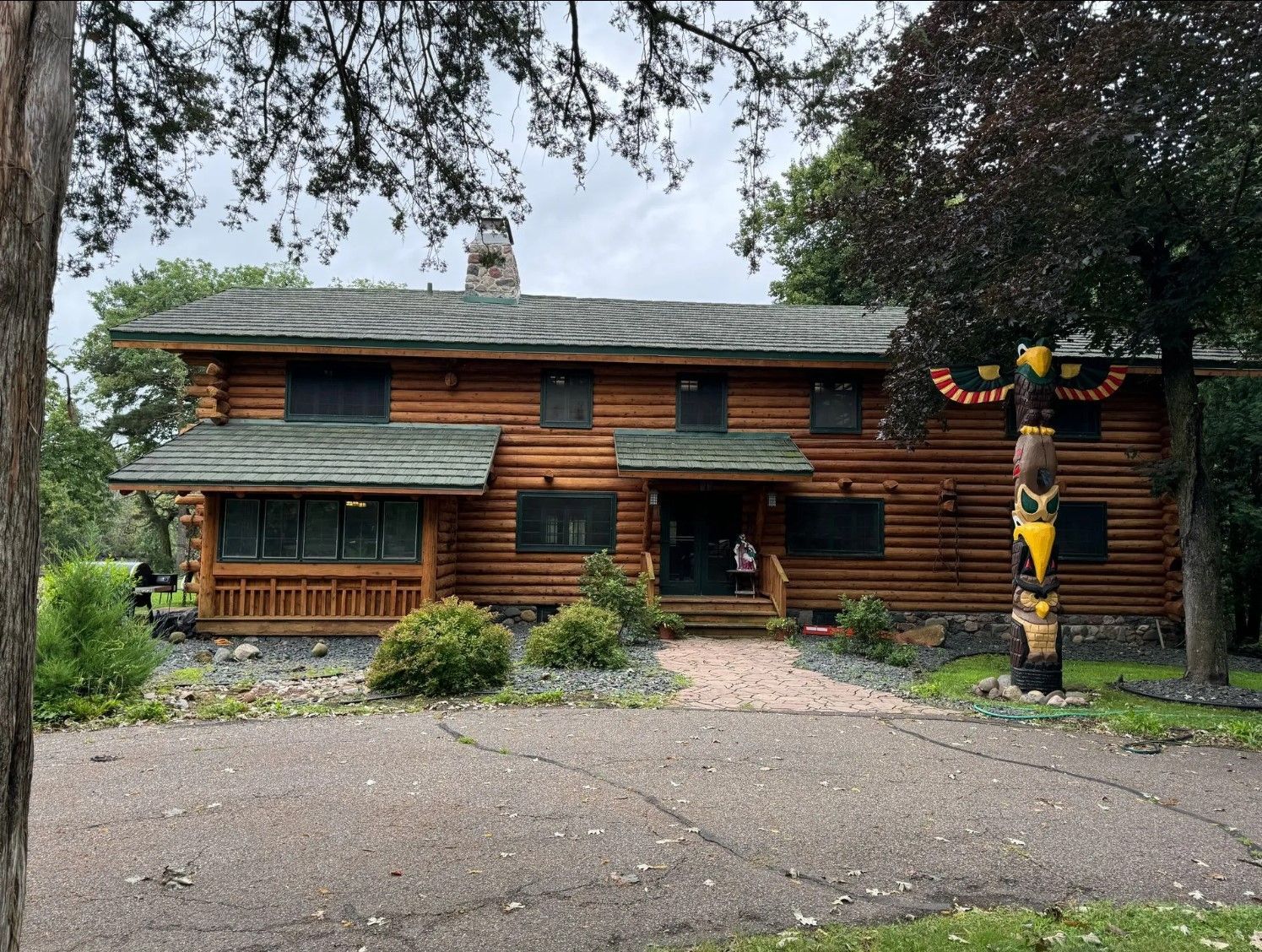 Two-story log cabin house with a driveway. Dark wood exterior, green roof. Tall tree on the right.