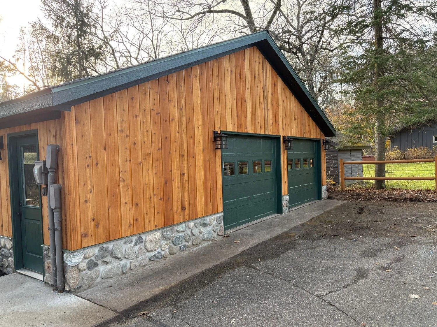 Wooden garage with green doors and roof; stone foundation. A driveway leads up to the doors.