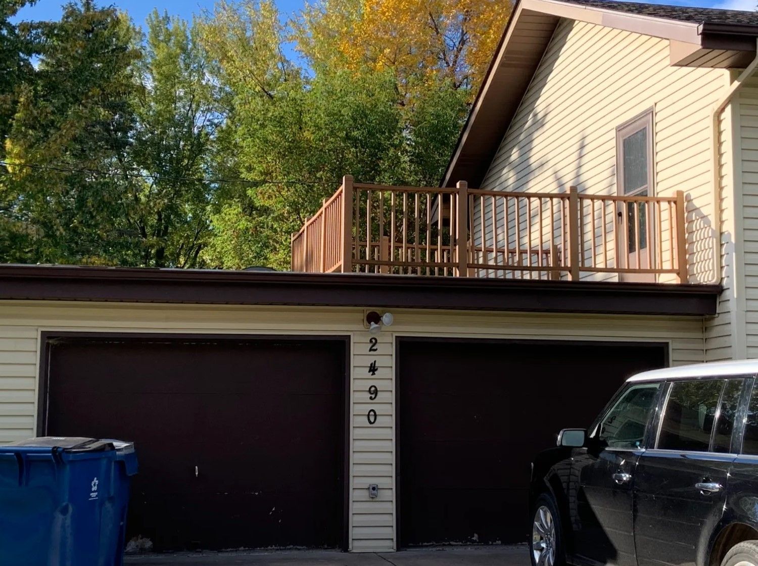 Two-car garage with a deck on top. A blue trash bin is to the left, and a car is parked on the right.