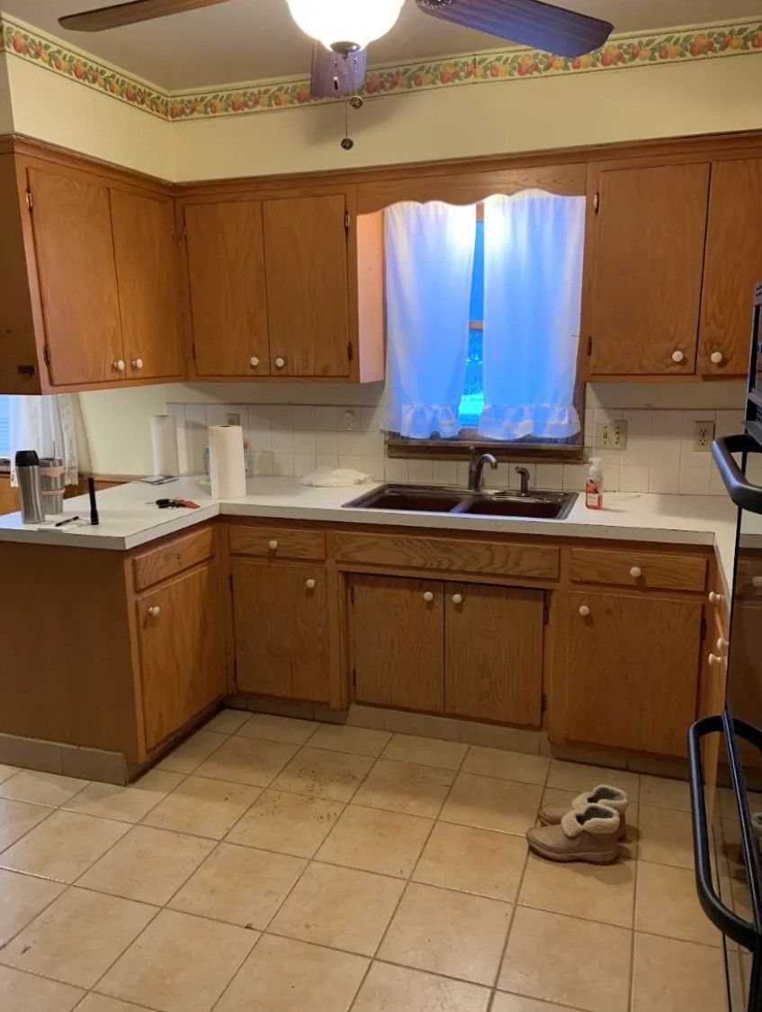 Kitchen with light brown cabinets, white countertops, and window with sheer curtain. Tan tile floor.