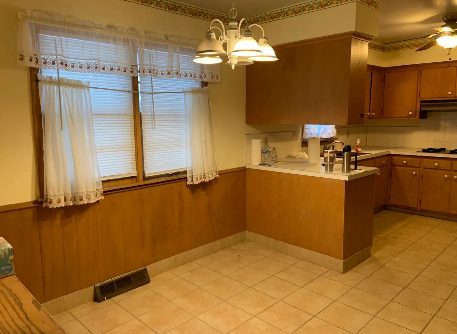 Kitchen with wood paneling, cabinets, and a breakfast bar. Windows with curtains and blinds. Beige tile floor.