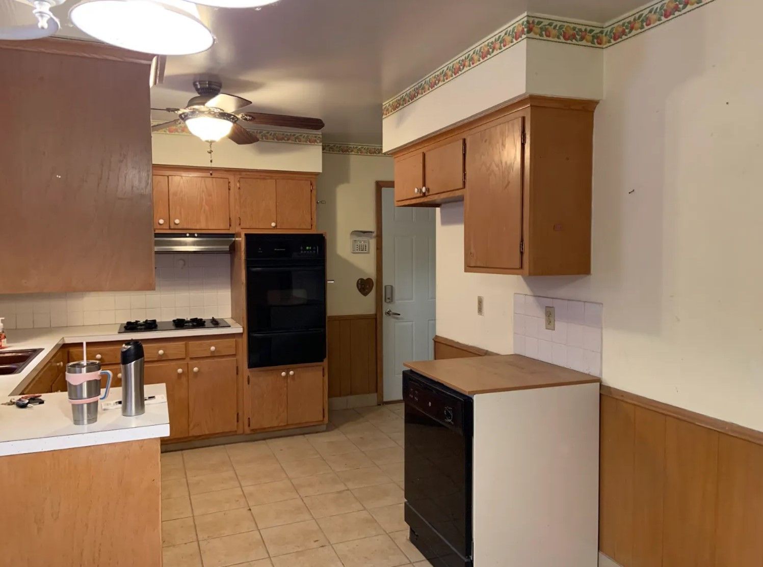 Kitchen with light wood cabinets, black oven, and dishwasher. Beige walls, floral border.