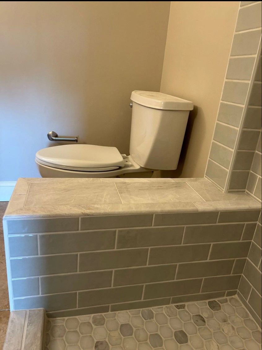 Toilet next to a gray tiled shower bench, with a stone shower floor. Beige walls.