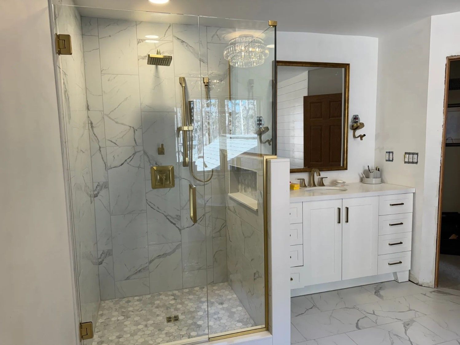 Modern bathroom with a glass shower, marble tile, and gold fixtures. White vanity and mirror.