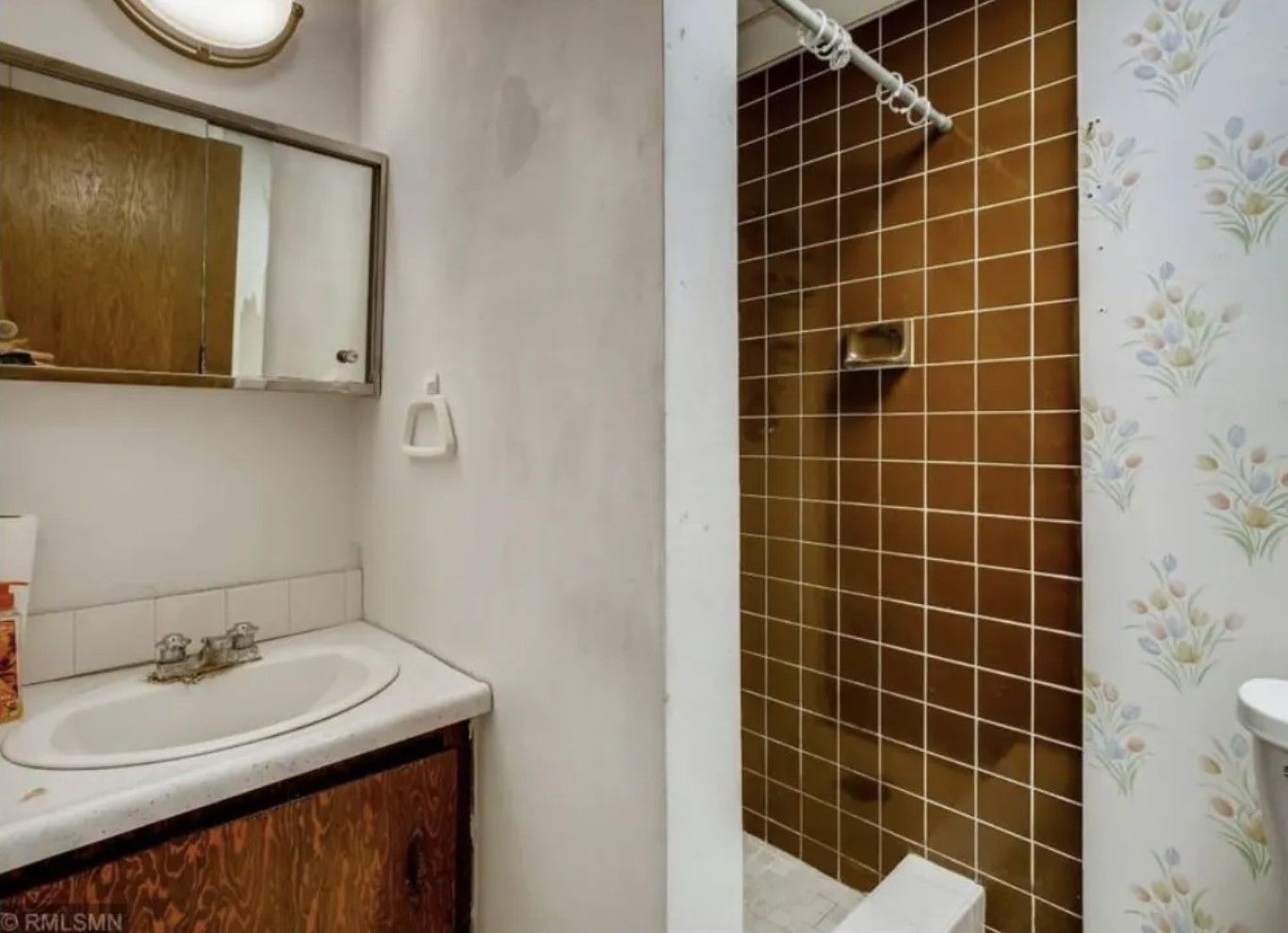 Small, dated bathroom with brown tiled shower, white sink, and floral wallpaper.