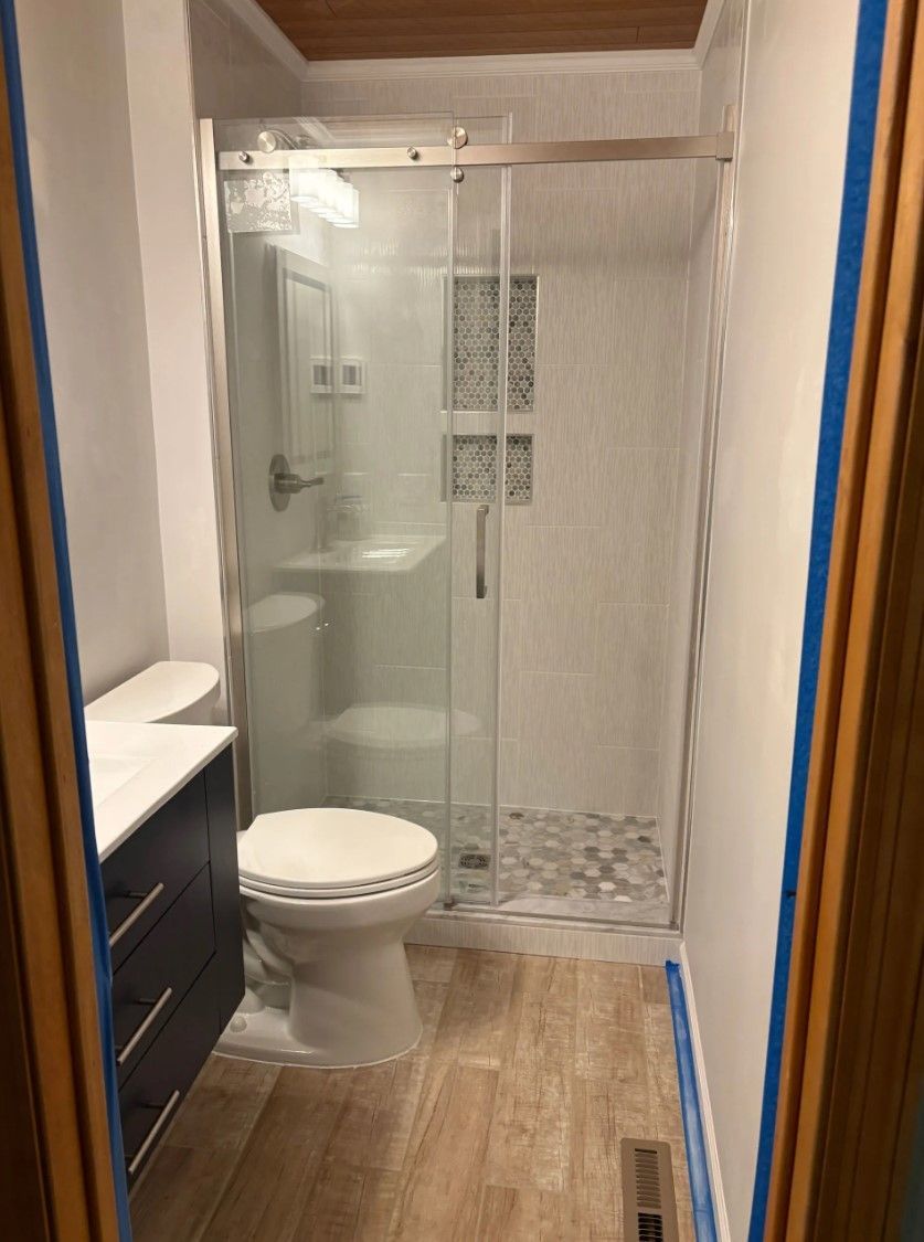 Bathroom interior with a shower, toilet, and vanity; light wood-look floor, white walls, and clear glass shower door.