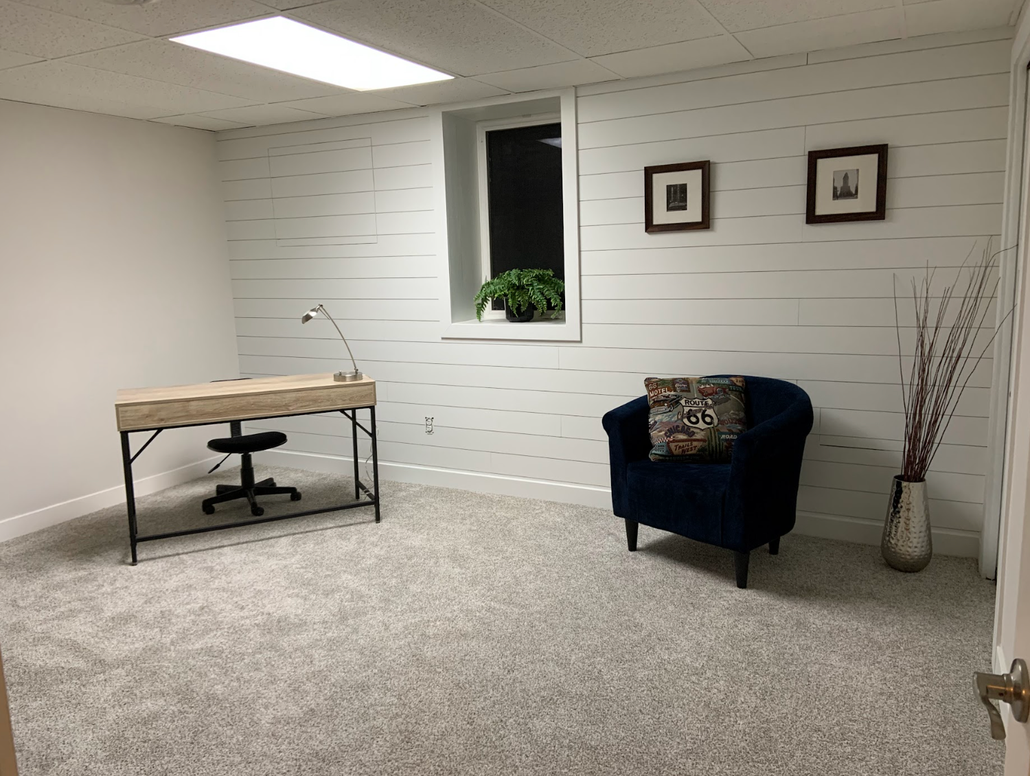 Home office with light gray carpet, desk, chair, blue armchair, window, and decorative vase.