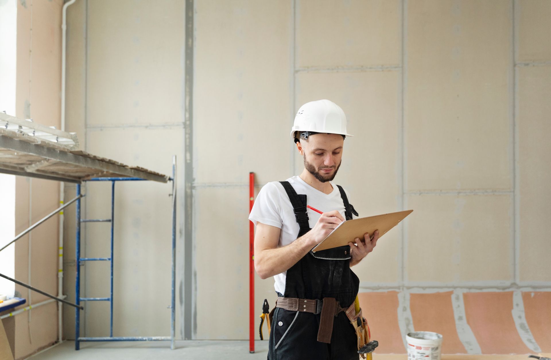 Construction worker in a white hard hat and overalls, writing on a clipboard in a room under renovation.
