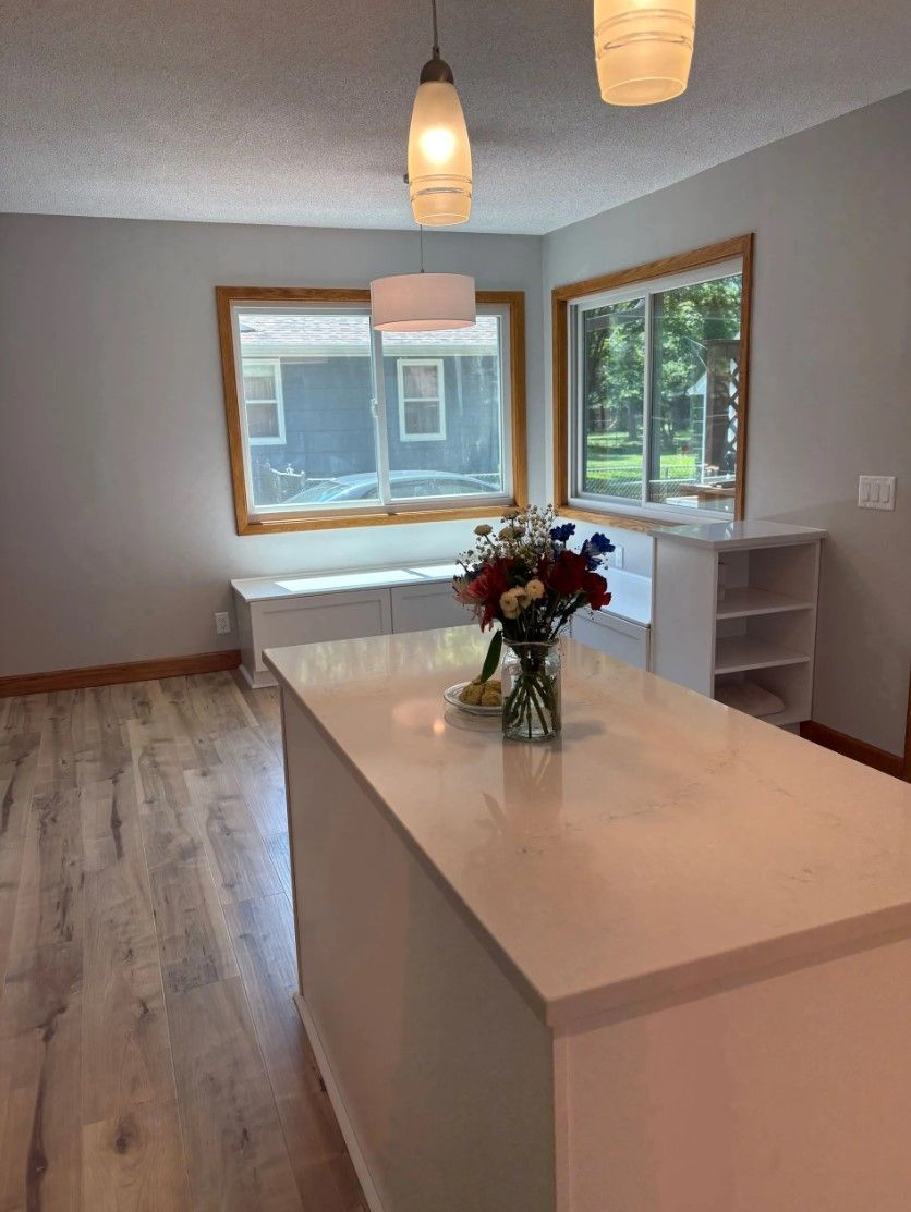 Kitchen with white island and countertops, wood trim around windows, and a vase of flowers.