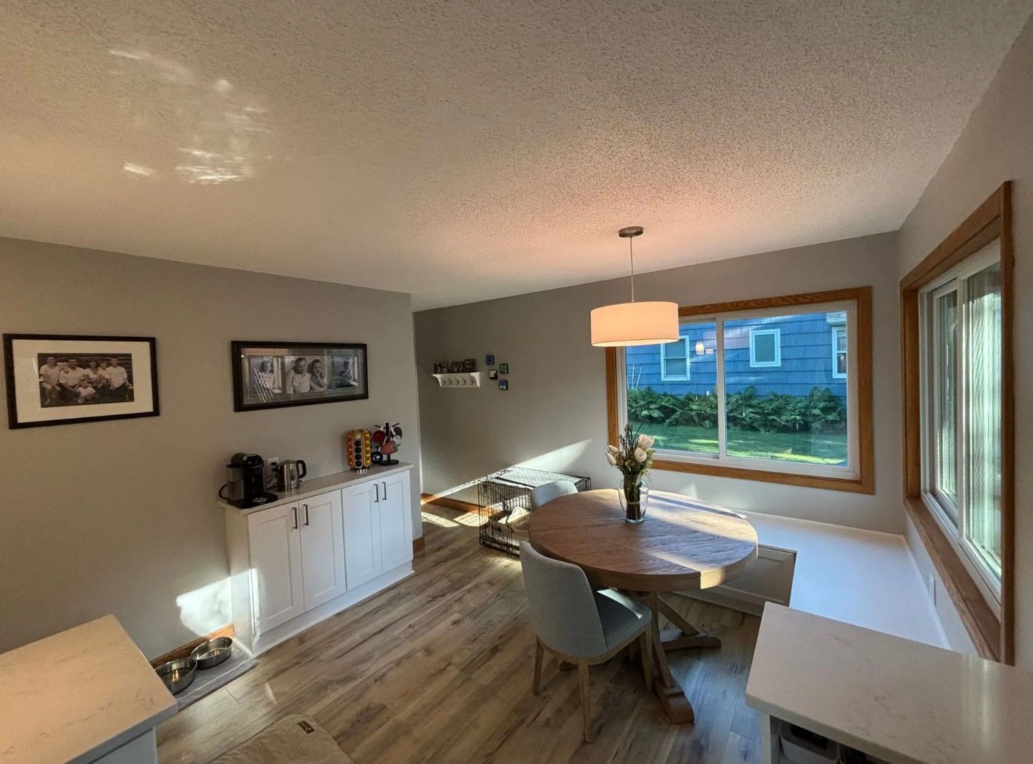 Dining room with a round wooden table, chairs, and large windows. Gray walls and white cabinets.
