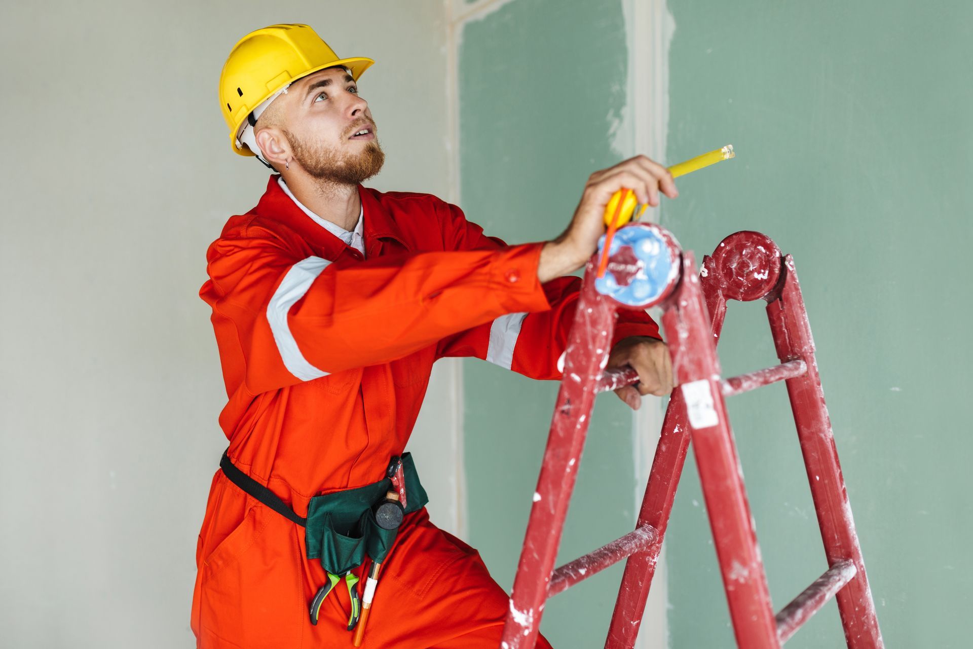 A man is sitting on a ladder measuring a wall with a tape measure.