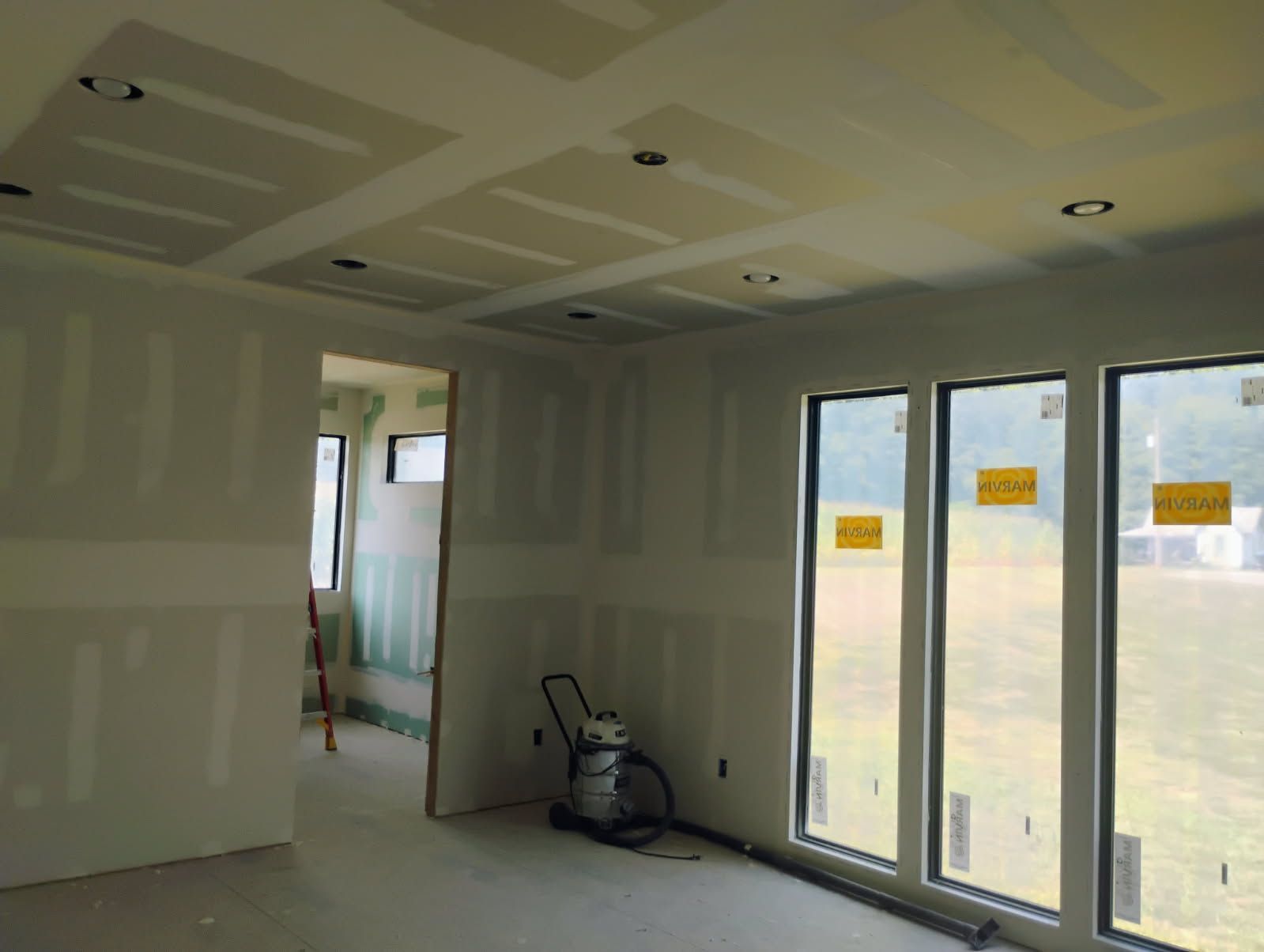 Interior of a room under construction, with drywall walls, ceiling, and large windows.