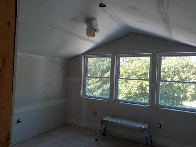 Interior view of a room under construction with drywall and three windows looking out to trees.