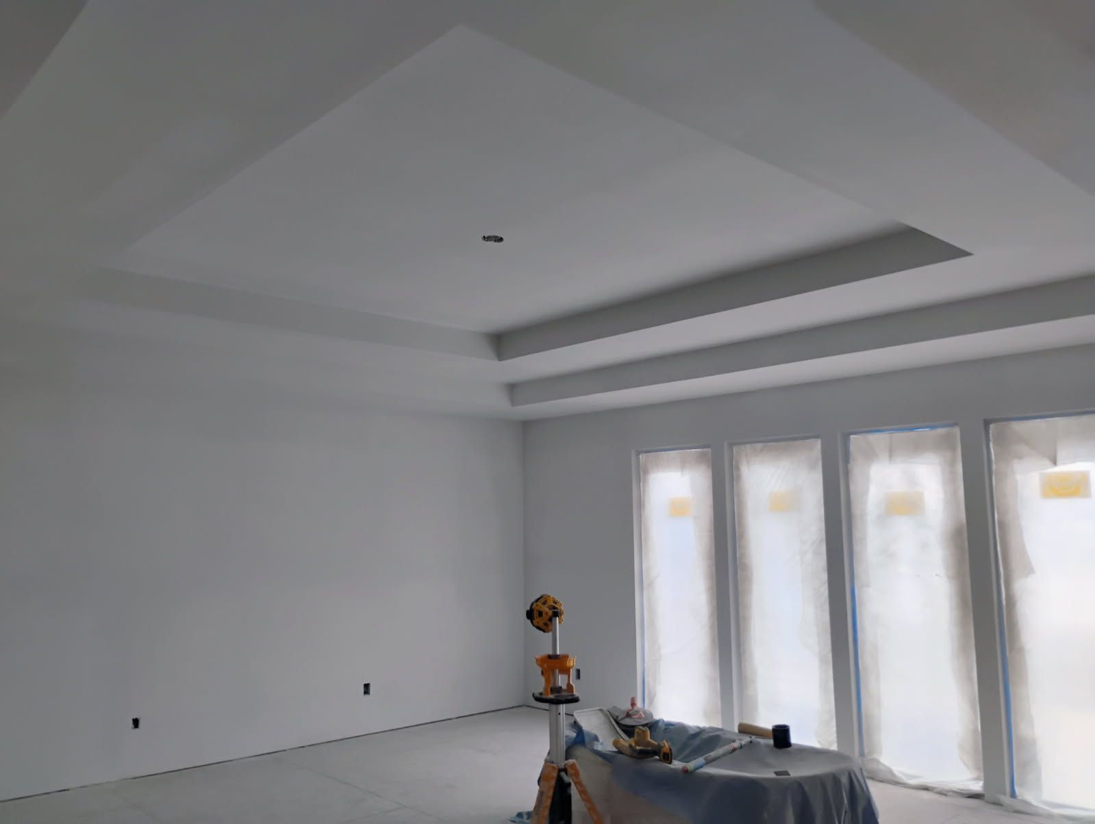 Empty room with white walls, multi-tiered ceiling, and large windows covered in plastic.