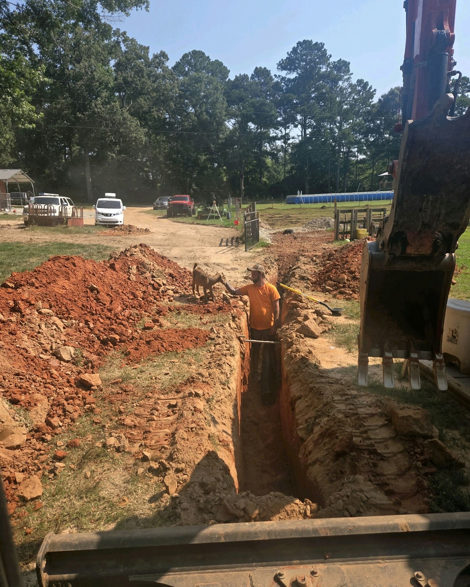 Construction trench with excavator, piles of red dirt, and vehicles near trees under a clear sky