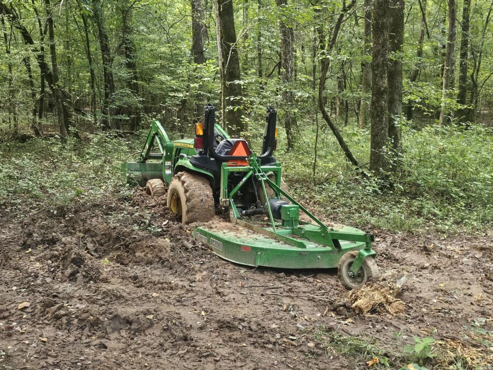 Green tractor mowing a muddy forest trail surrounded by trees