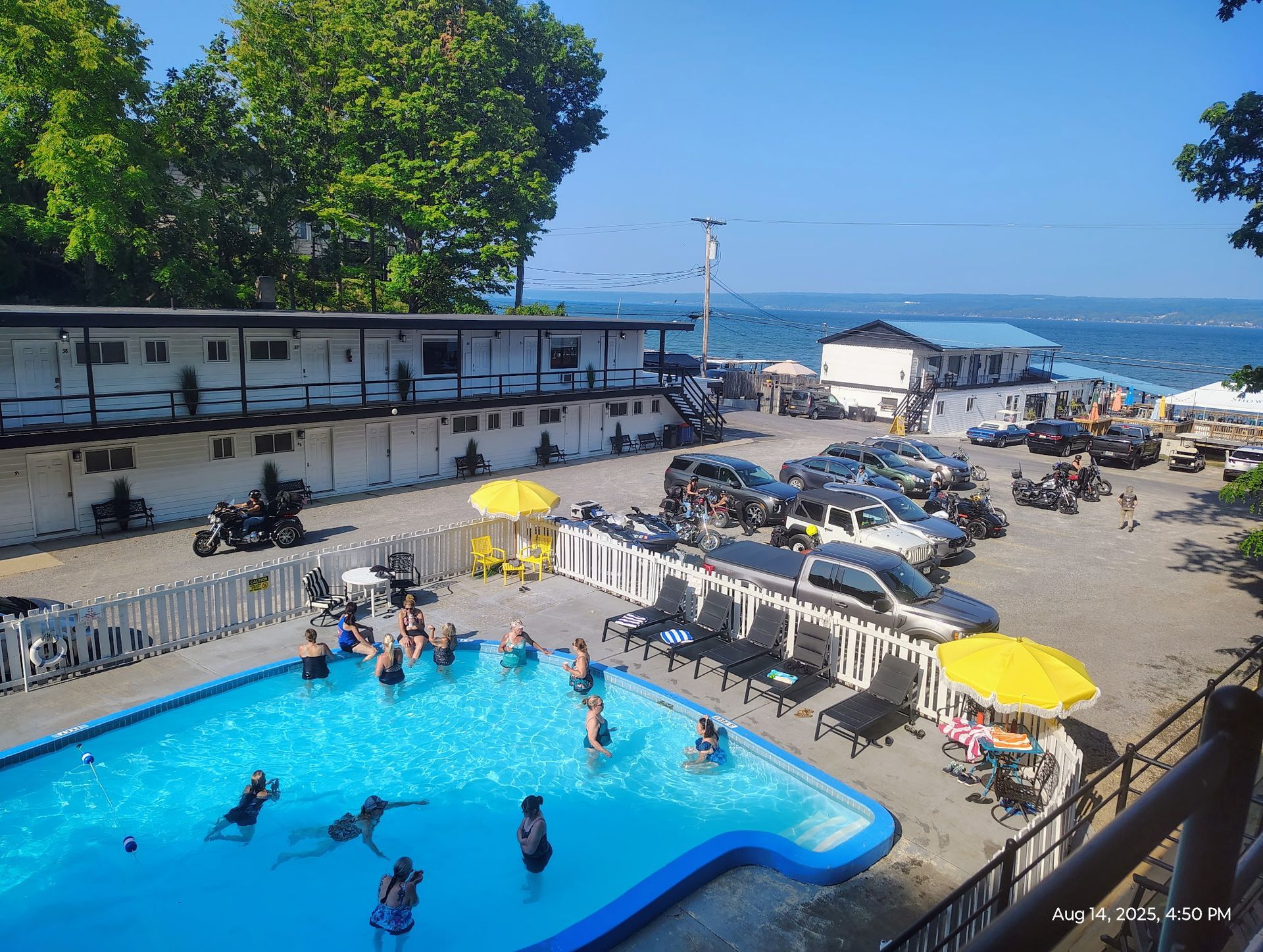 Pool with building in the background. Blue water and sky. Wooden porch, trees and white chairs.