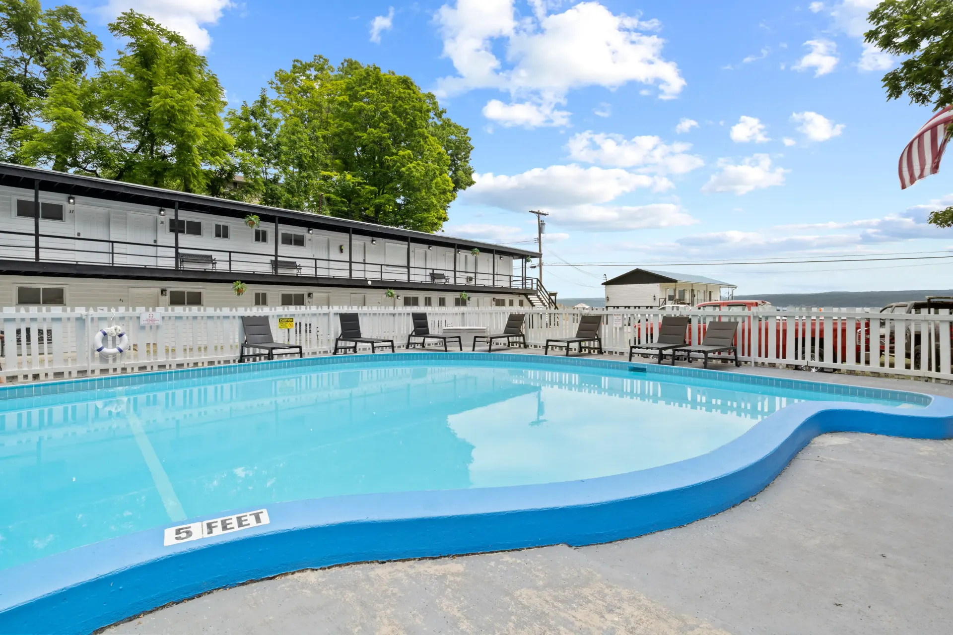 An outdoor swimming pool with bright blue water and a painted deck, set against a building with a white fence and trees.