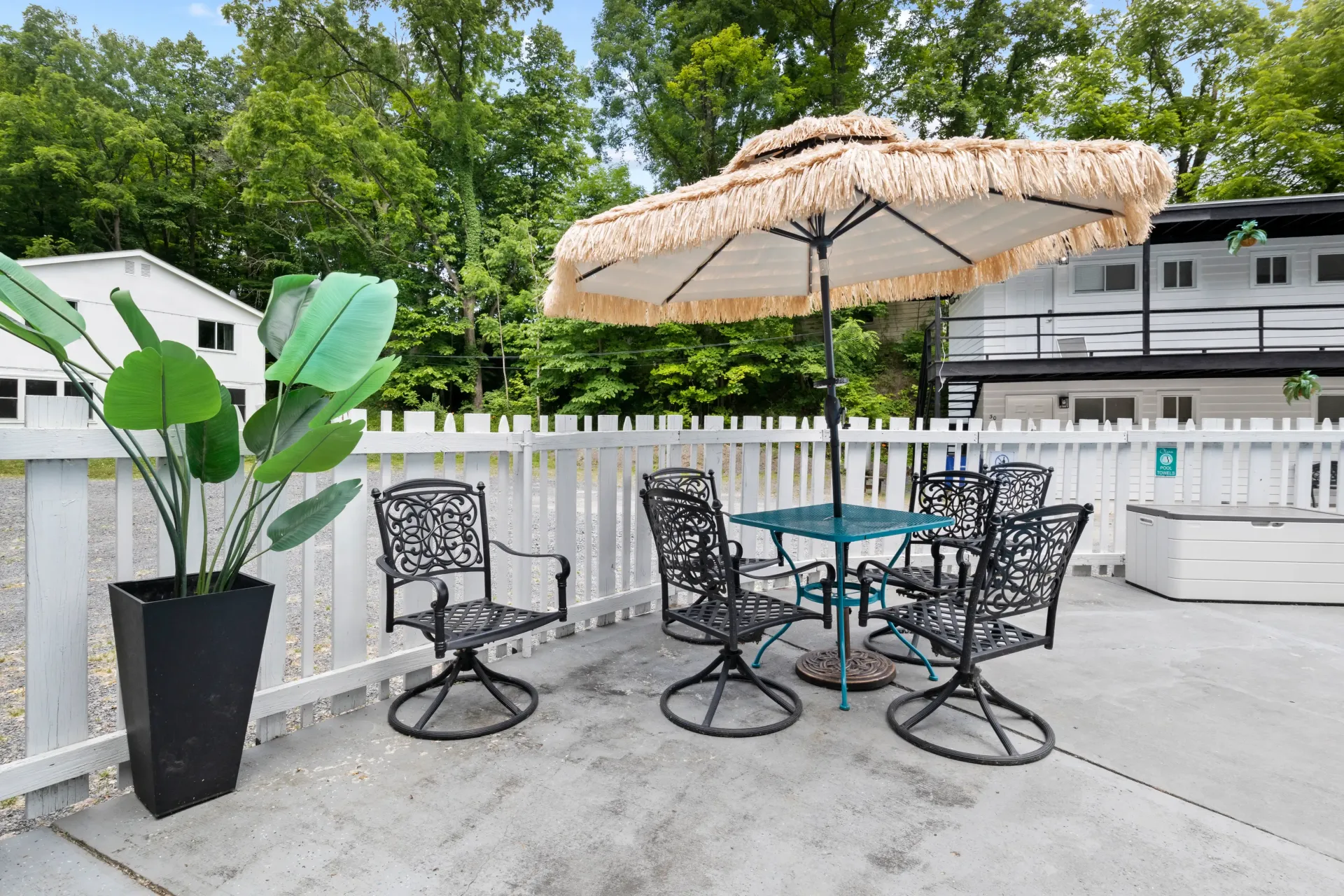 Outdoor patio with a teal umbrella, four black metal chairs, a matching table, and a large potted plant on concrete.