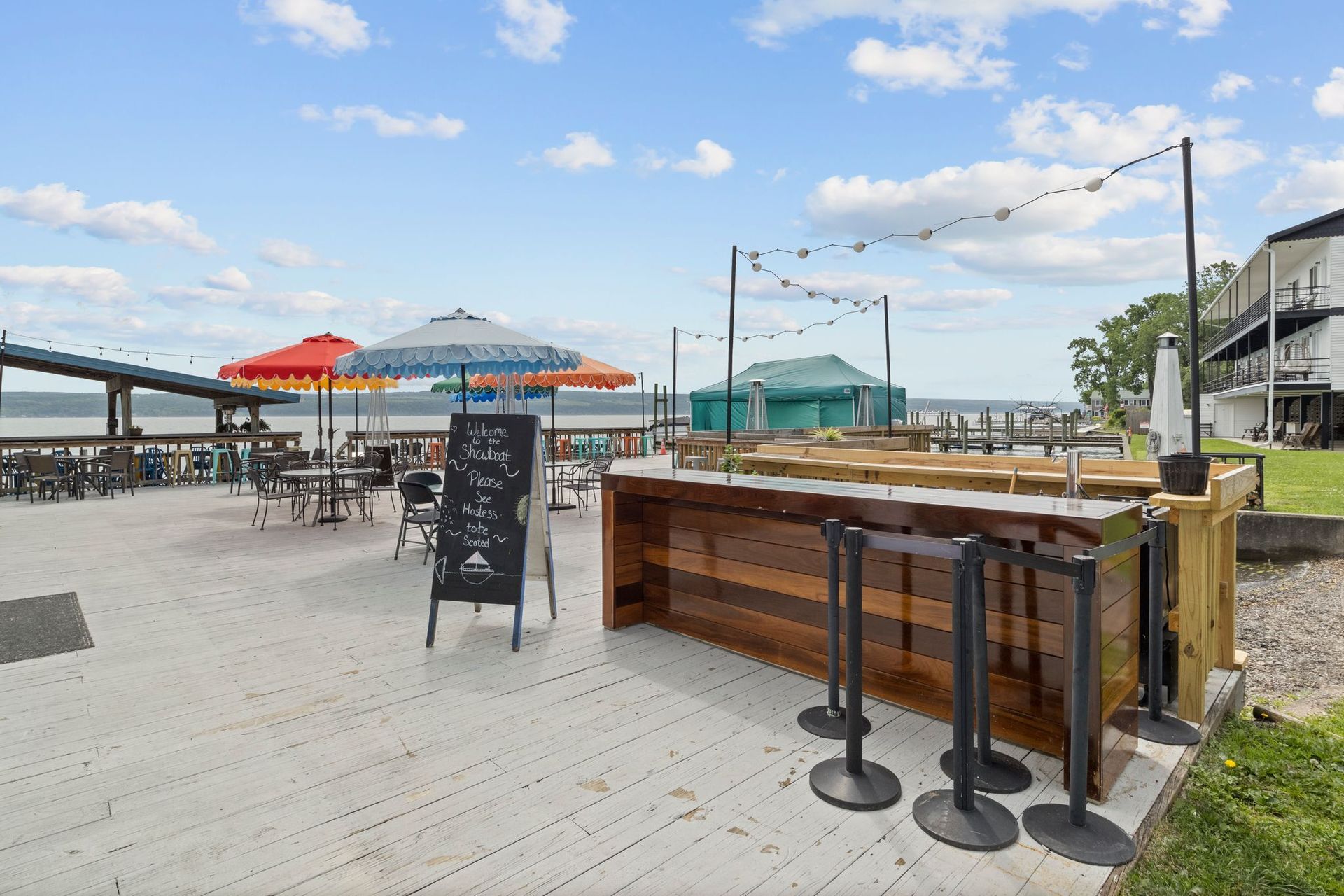 An outdoor patio bar by a waterfront with colorful umbrellas, a chalkboard menu, and a wooden counter under string lights.