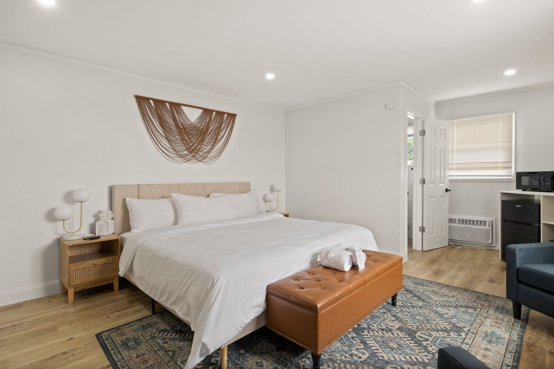 A modern hotel bedroom with a king bed, tan bench, macramé wall art, and light wood floors, viewed from the doorway.