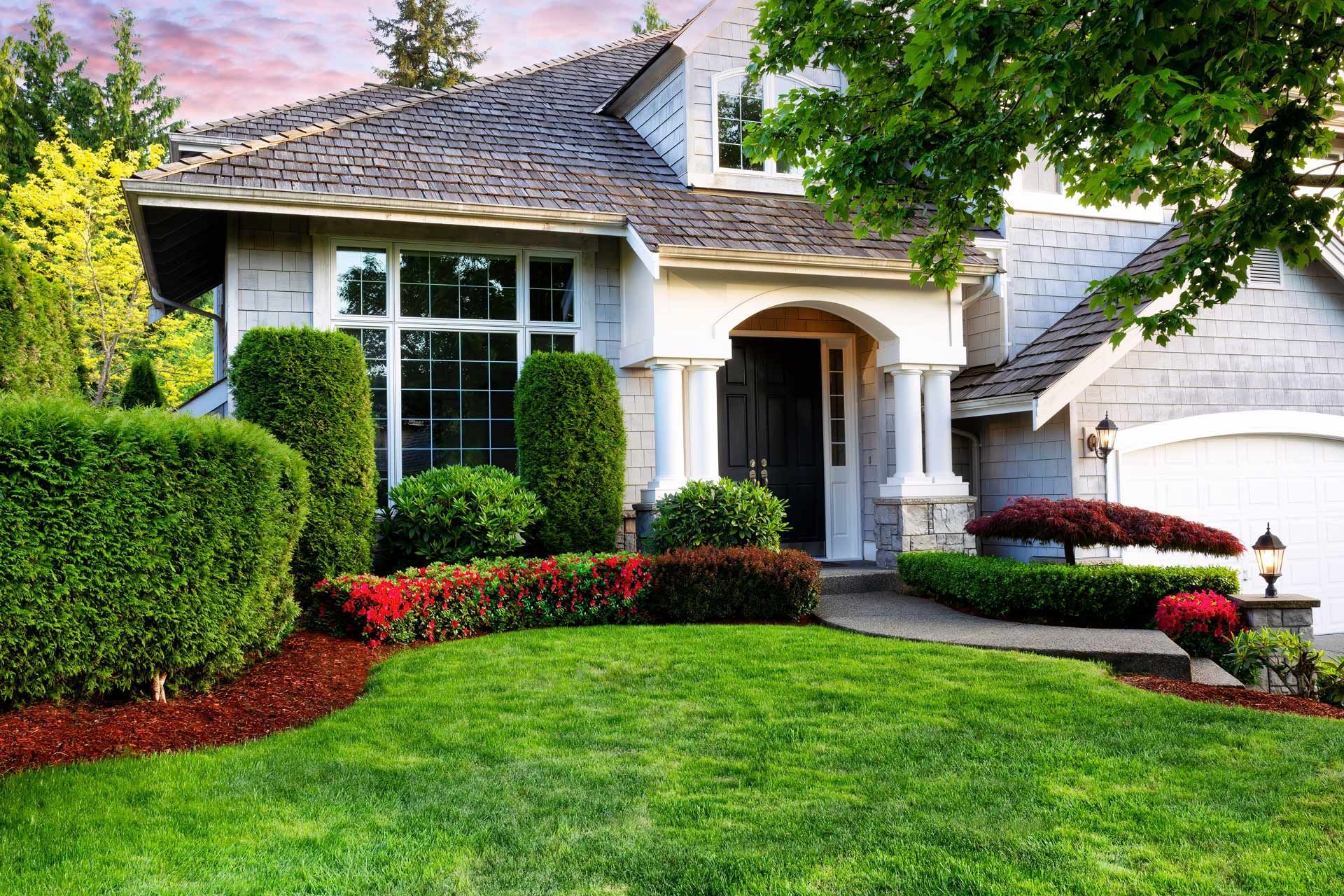 A suburban home with a green lawn, neatly trimmed hedges, red flowers, and a covered front entryway.