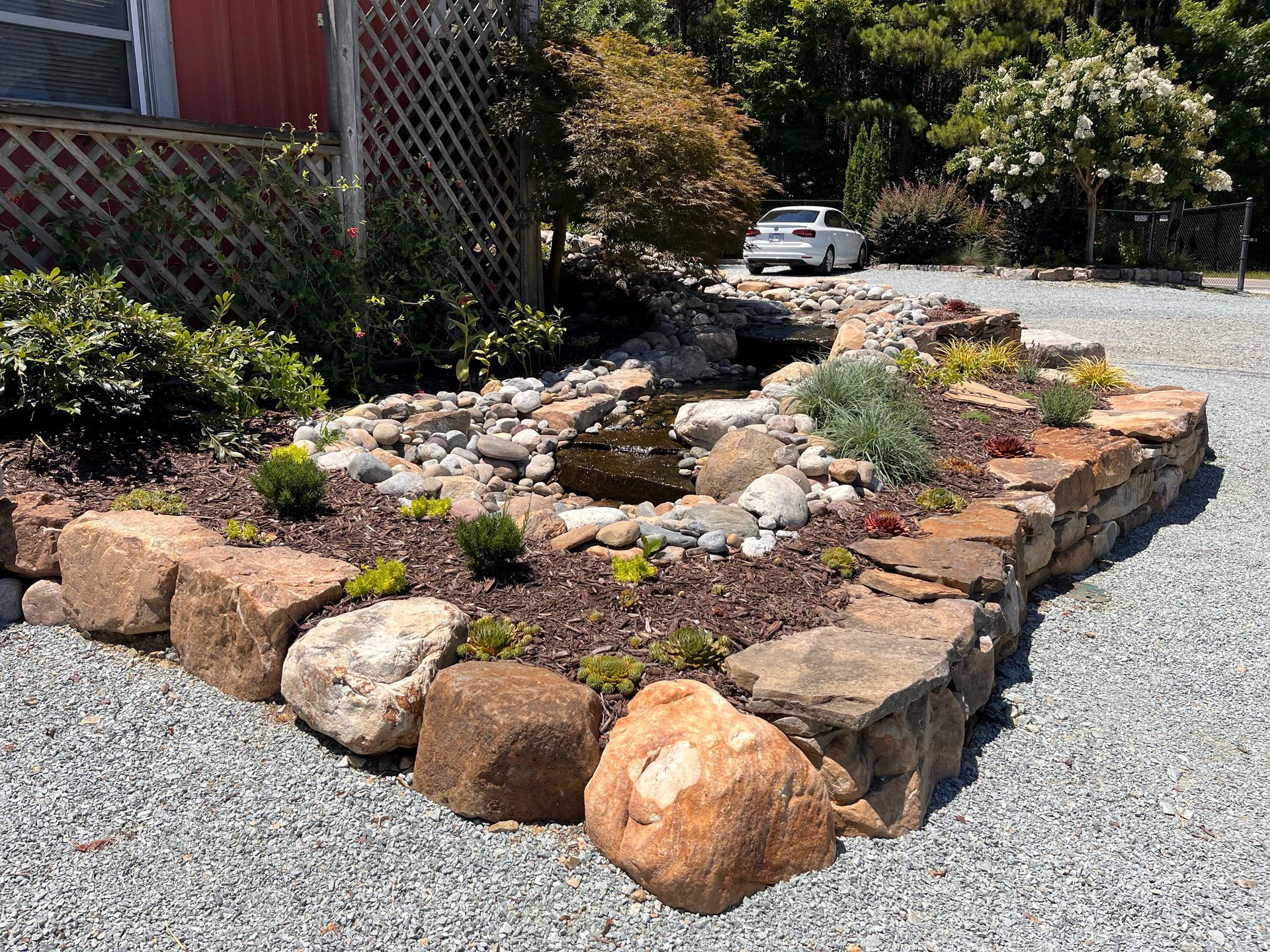 Stone-walled garden bed with a small pond and waterfall, gravel surrounding. A white car in the background.