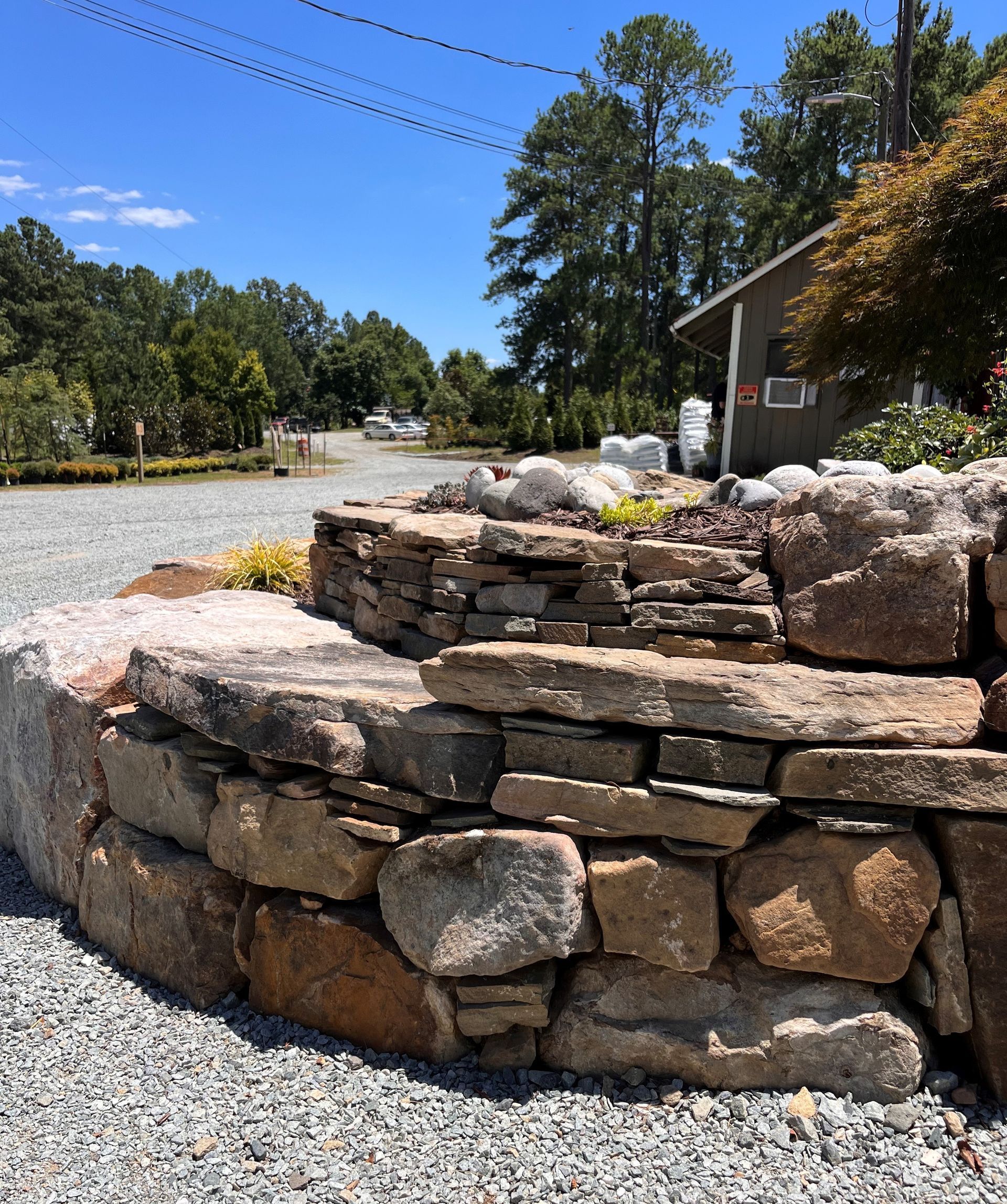 Stone retaining wall with layers and a gravel driveway in front of a building and trees.